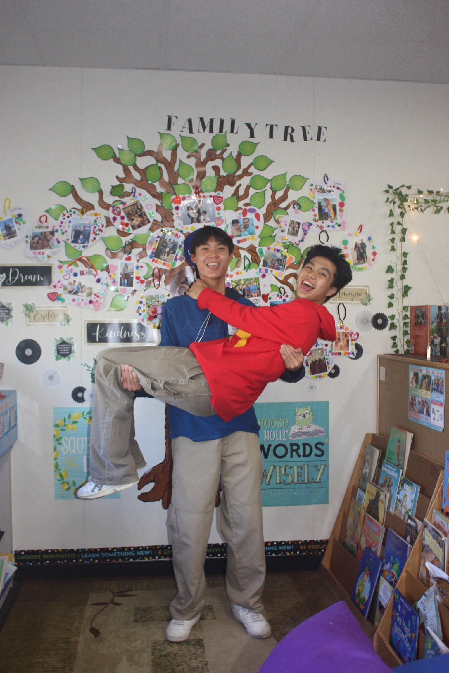 Two young men are standing in a classroom with a wall display of a family tree and photos behind them. One man is holding the other in his arms, and both are smiling. There are bookshelves and educational posters in the background.