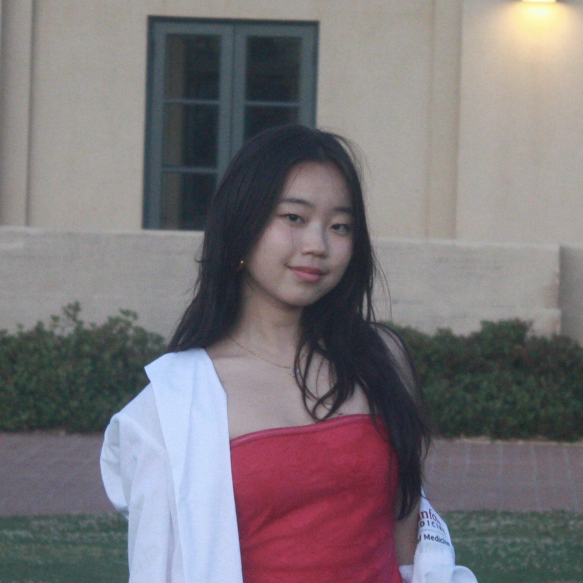 A young woman with long black hair standing outdoors at dusk, smiling slightly, wearing a red tube top and a white jacket draped over her shoulders, with a building and window in the background.
