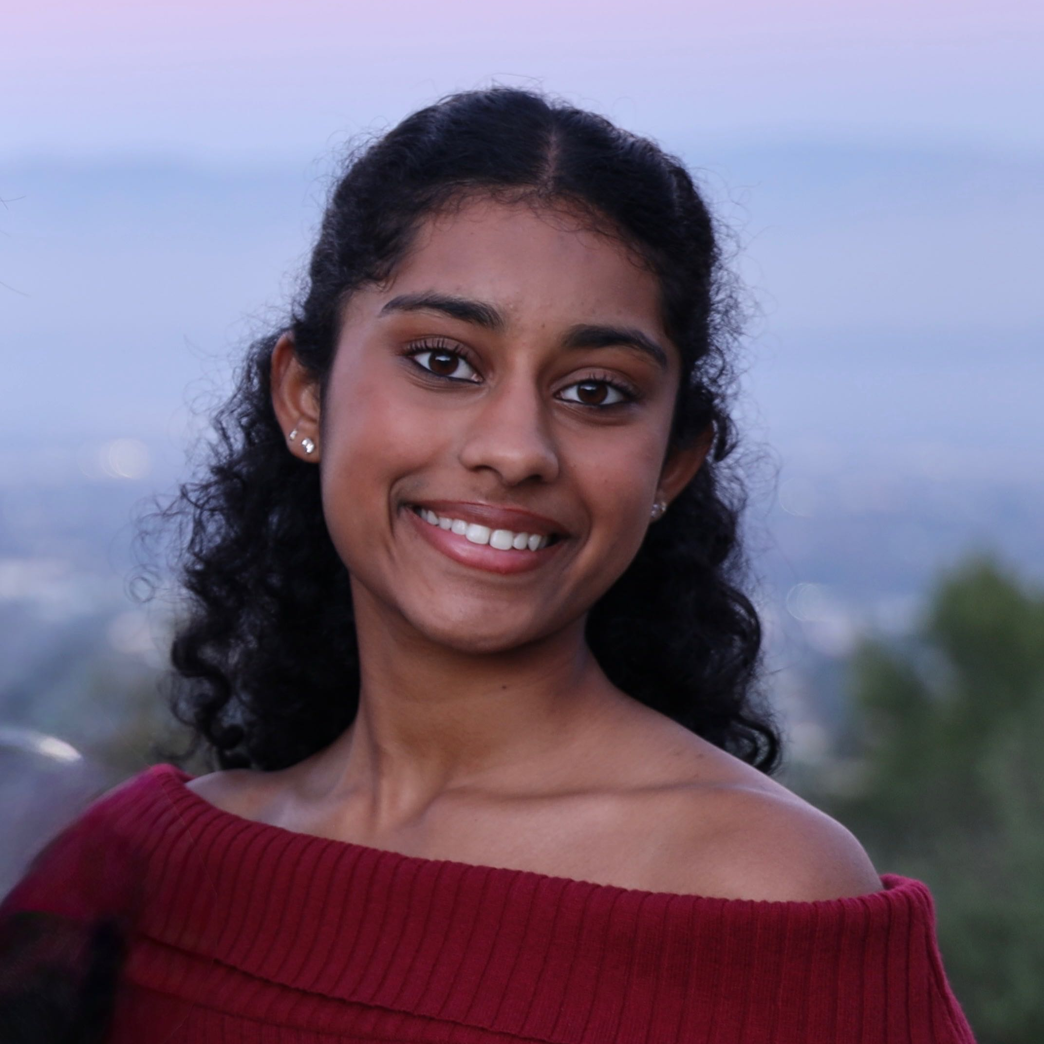 Close-up of a smiling woman with dark curly hair, wearing earrings and a red sweater, outdoors during dusk or dawn with a blurred landscape background.