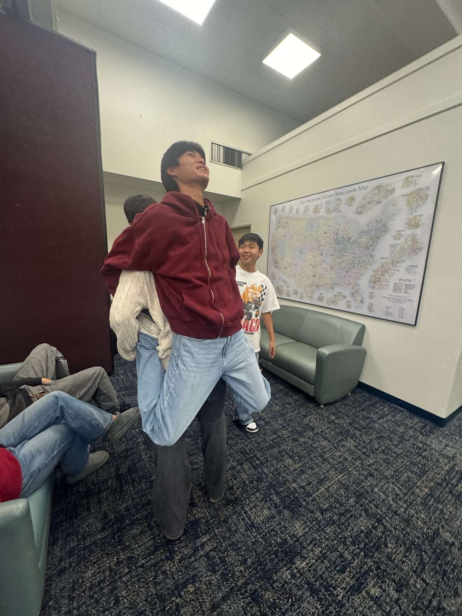 Two young men are standing and smiling in a room with a large map of the United States on the wall. People are sitting or lying on the floor nearby.
