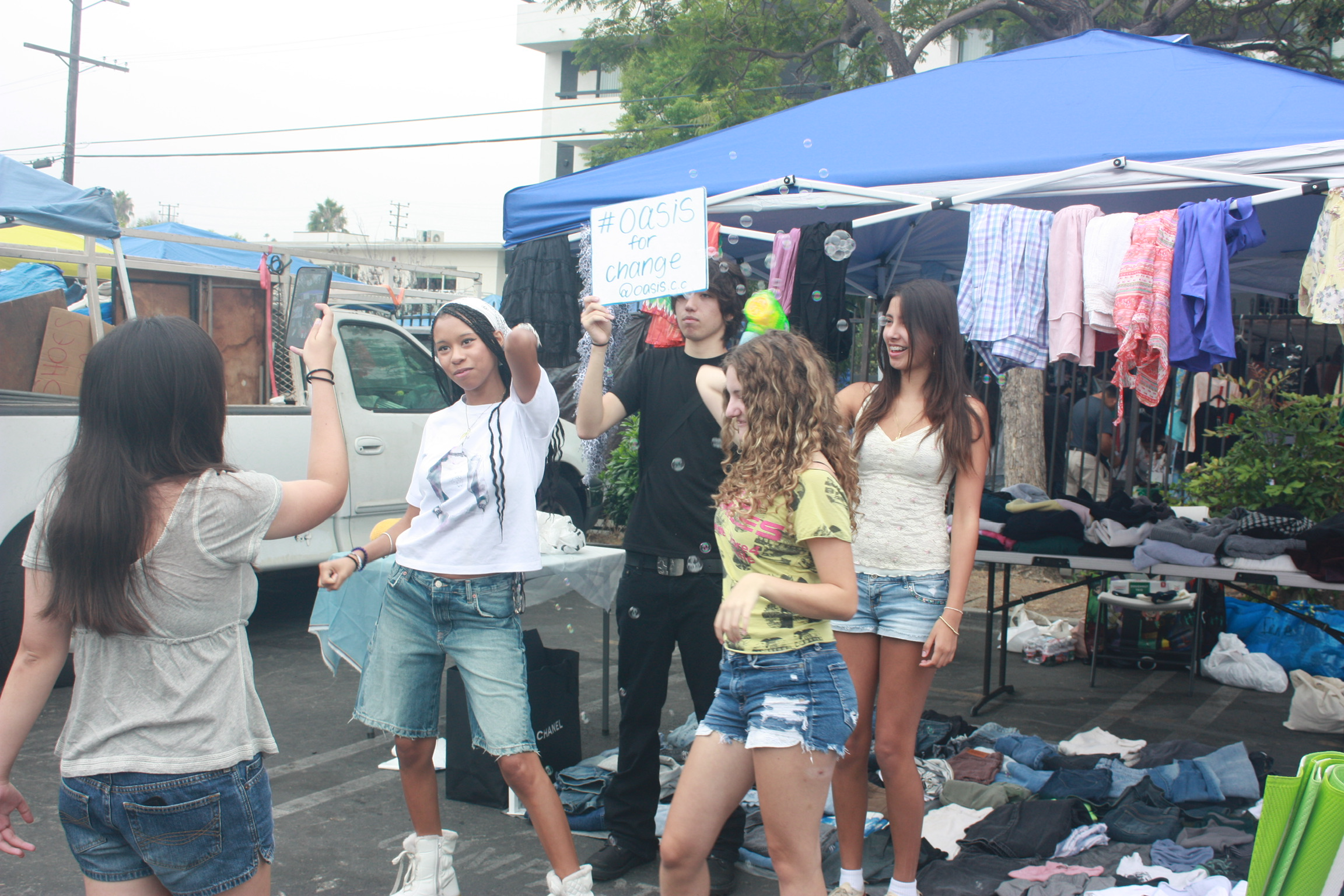 Group of people at a yard sale with clothing on tables and hanging on racks under tents, and a sign reading '#Oasis for change' held by a person in black.