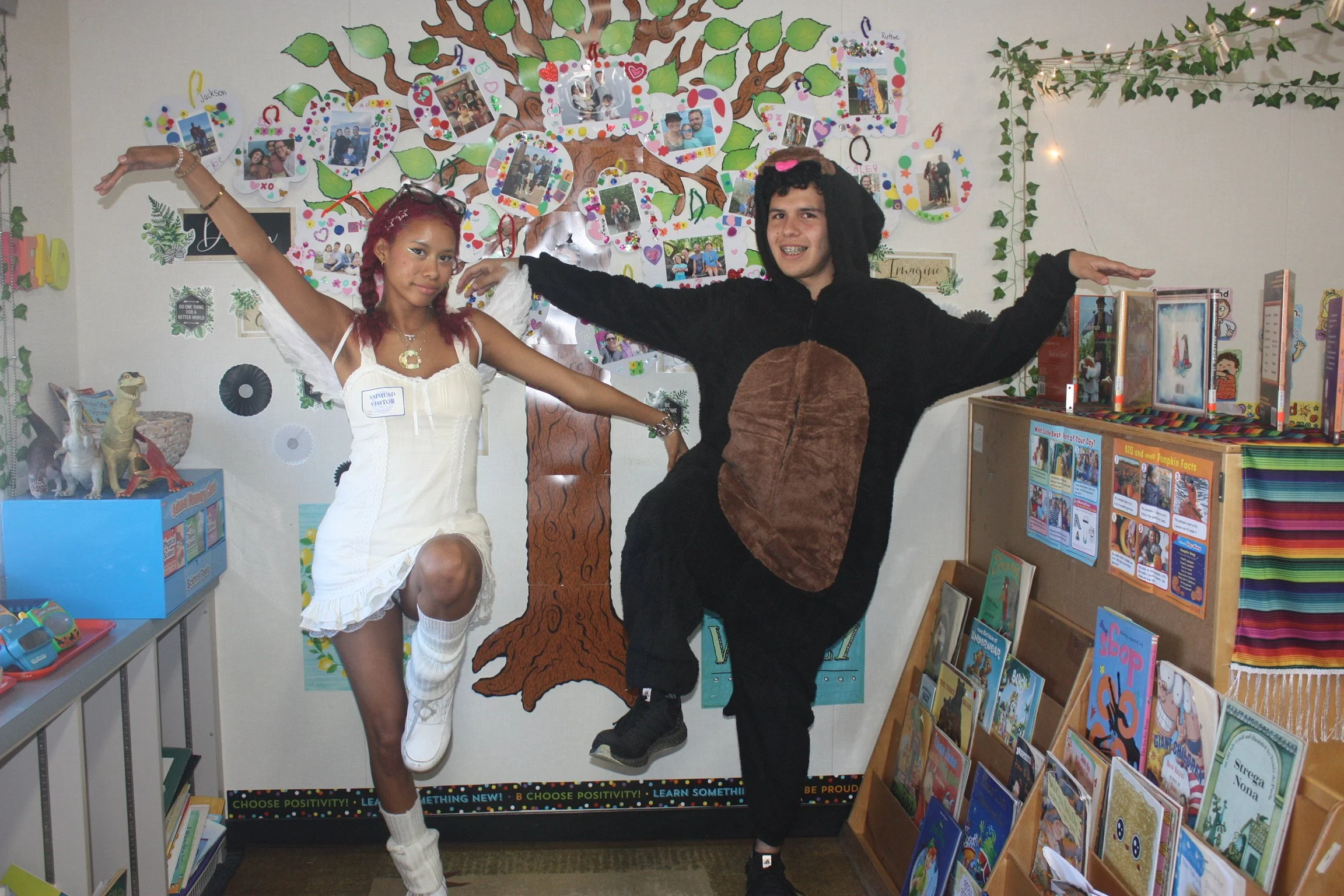 Two young people are dressed in costumes, one as a soccer player and the other as a bear, and are posing with arms outstretched in front of a decorated wall with photos and artwork. The girl has red hair and is wearing a white dress, while the boy ha