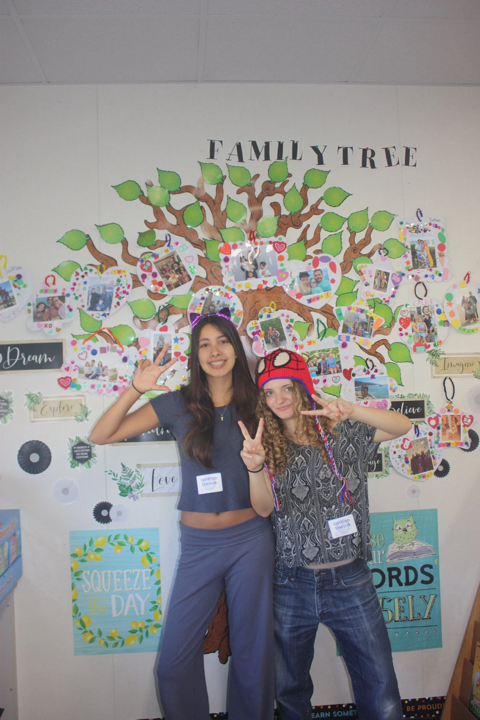 Two young women standing in front of a decorated wall with a tree and photos, smiling and making peace signs.