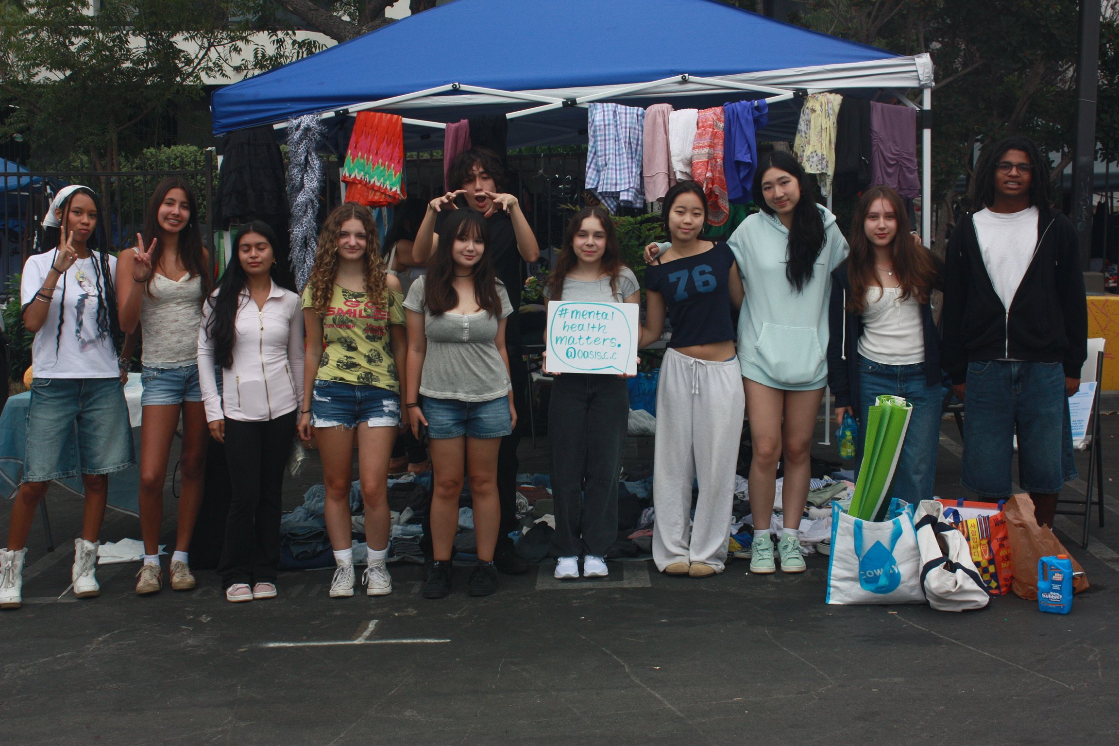 Group of teenagers standing together at a community event with a clothing donation booth in the background, some holding a sign that reads '#mental health matters.'