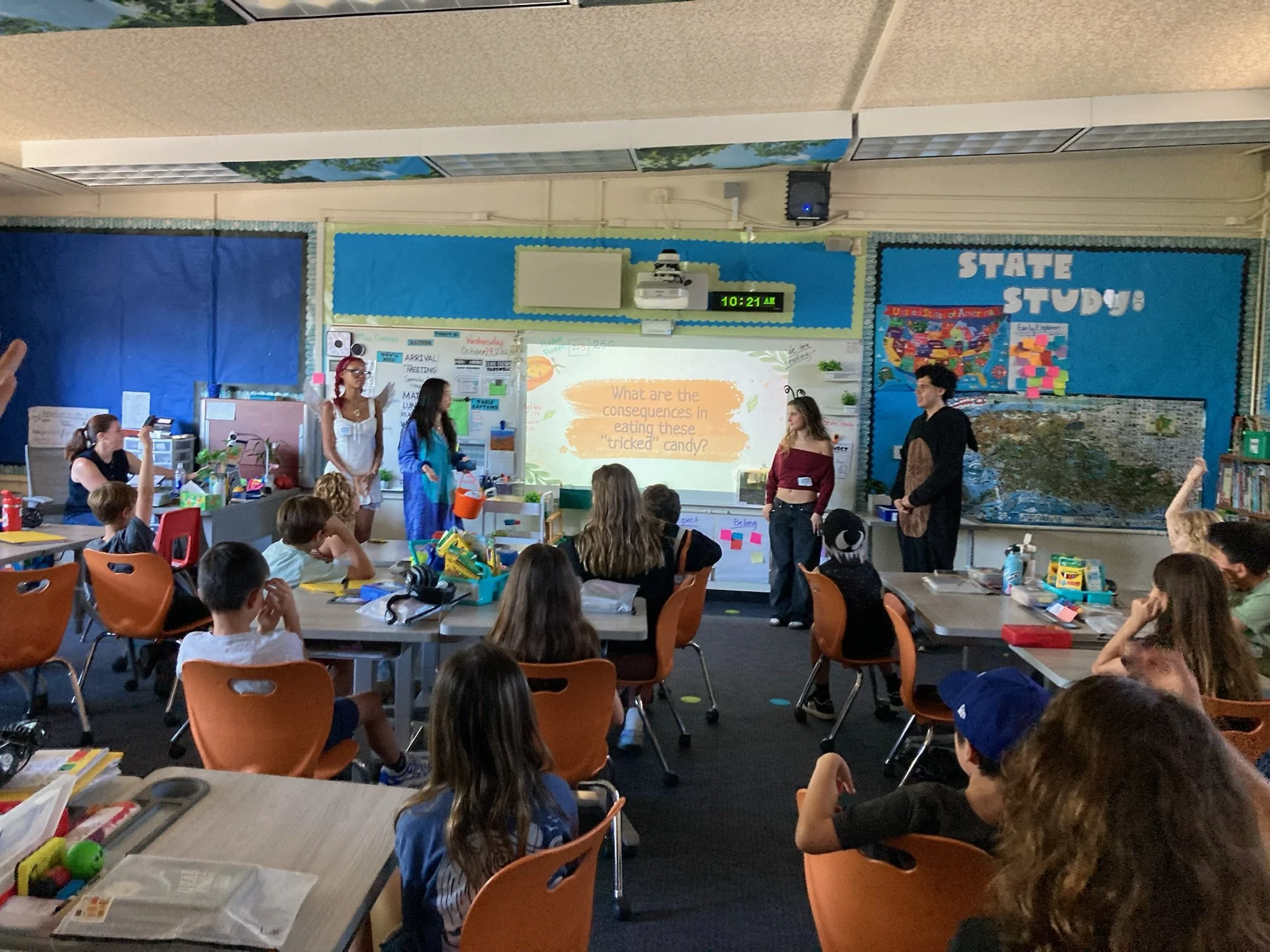A classroom with students sitting at desks and a group of students standing at the front, some dressed in costumes, with a whiteboard displaying a question about the consequences of eating tricked candies. The classroom has colorful decorations and m
