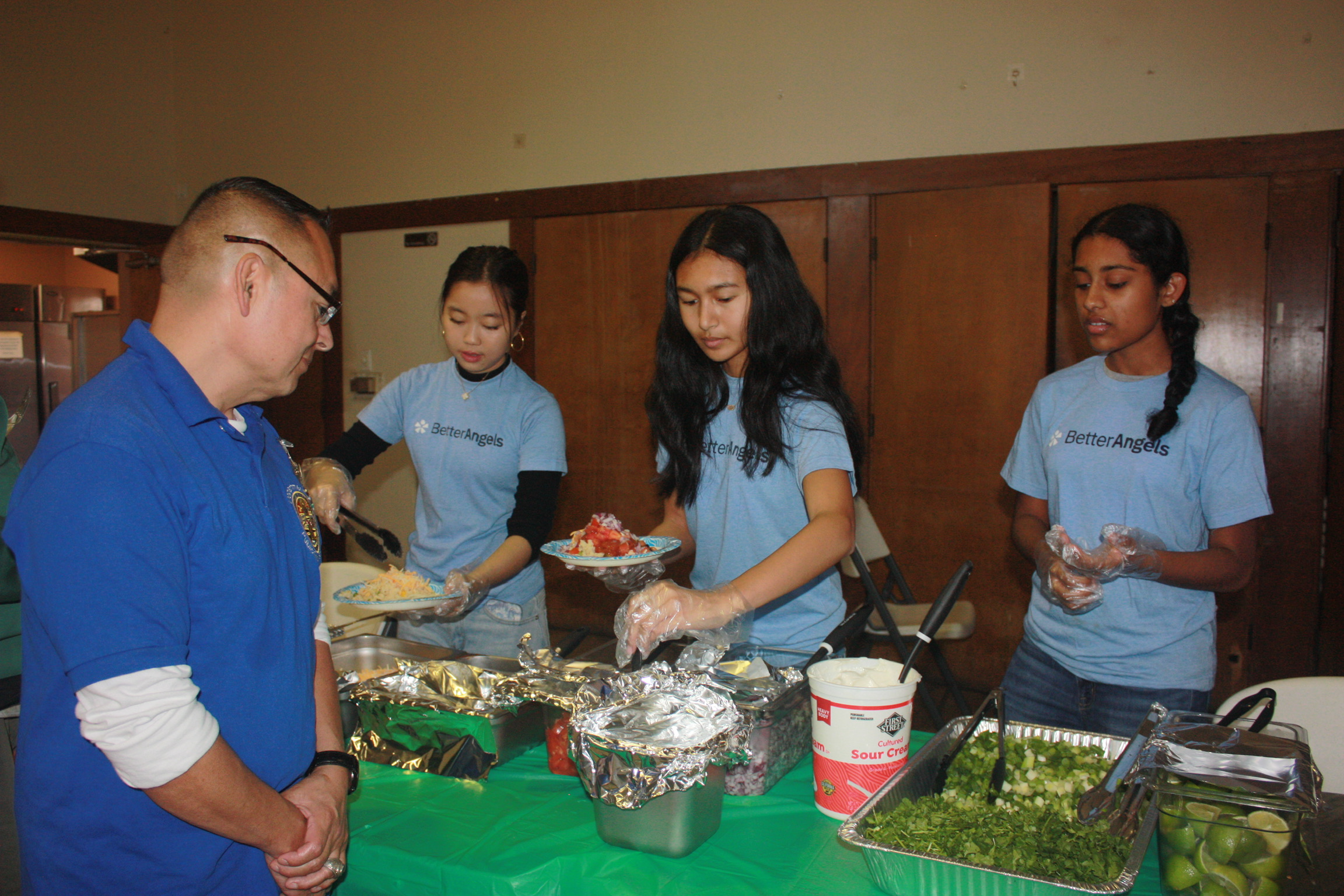Food service event with three young women in light blue t-shirts serving a variety of dishes, including salad and dessert, at a table covered with a green tablecloth.