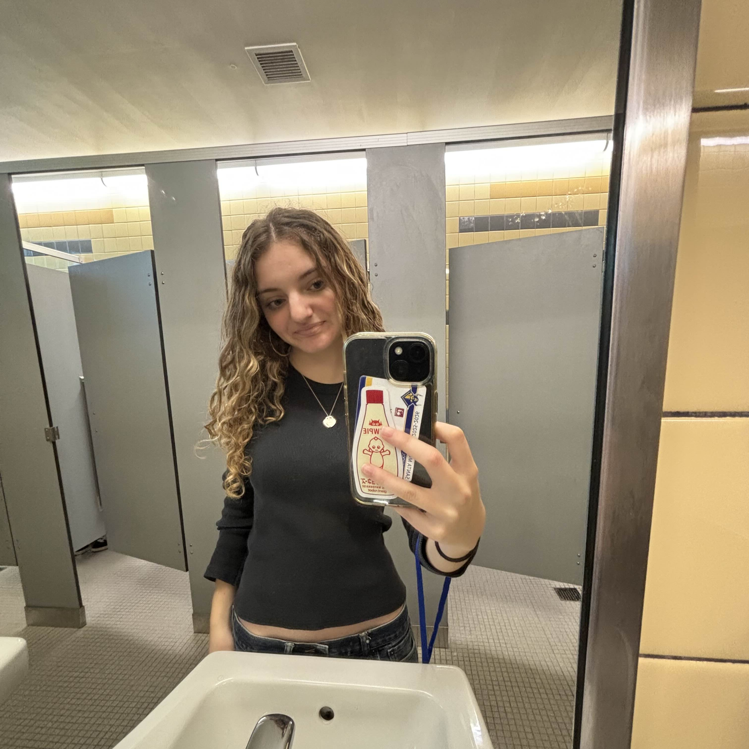 A young woman with long curly hair taking a selfie in a bathroom mirror. She is wearing a black top and is holding her phone with a case featuring cartoon characters. The bathroom has yellow and black tiled walls, a white sink, and gray stall dividers.
