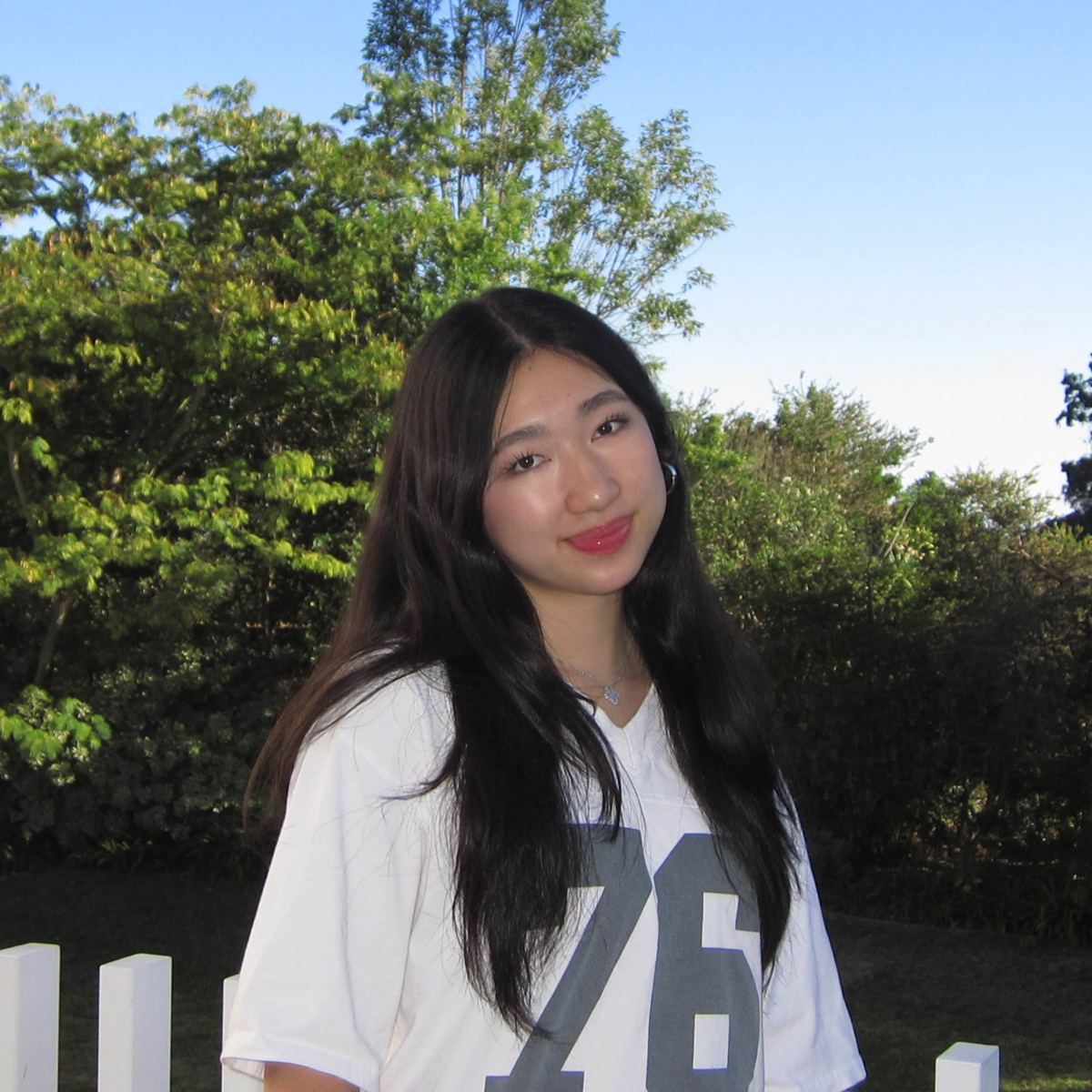 A young woman with long black hair, wearing a white sports jersey with the number 76, standing outdoors near a white fence, with green trees and a clear blue sky in the background.