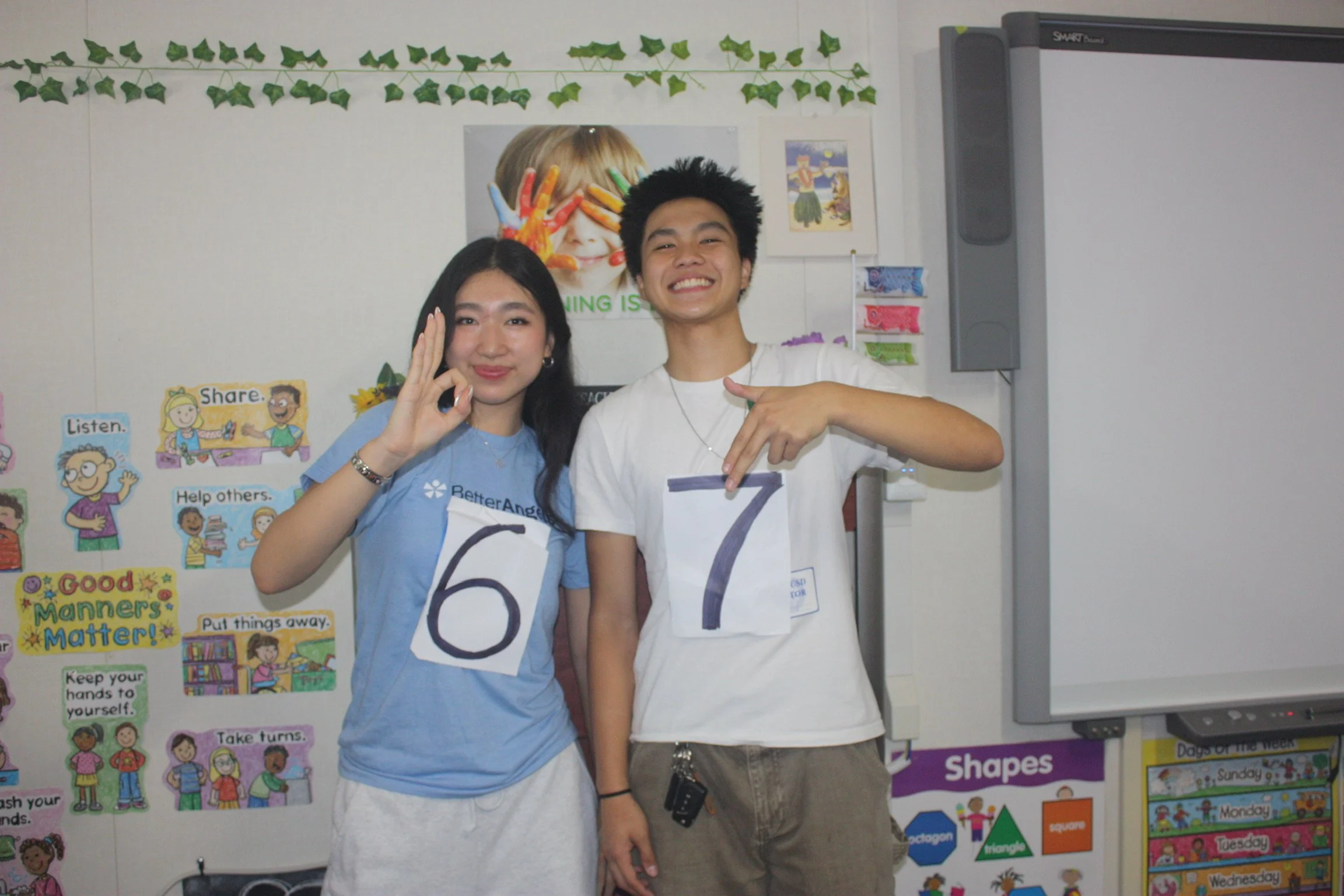 Two teenagers standing in front of a classroom bulletin board, both smiling and holding signs with the numbers 6 and 7. The girl is wearing a blue shirt with a Better Angels logo, and the boy is wearing a white shirt. They are making hand gestures an