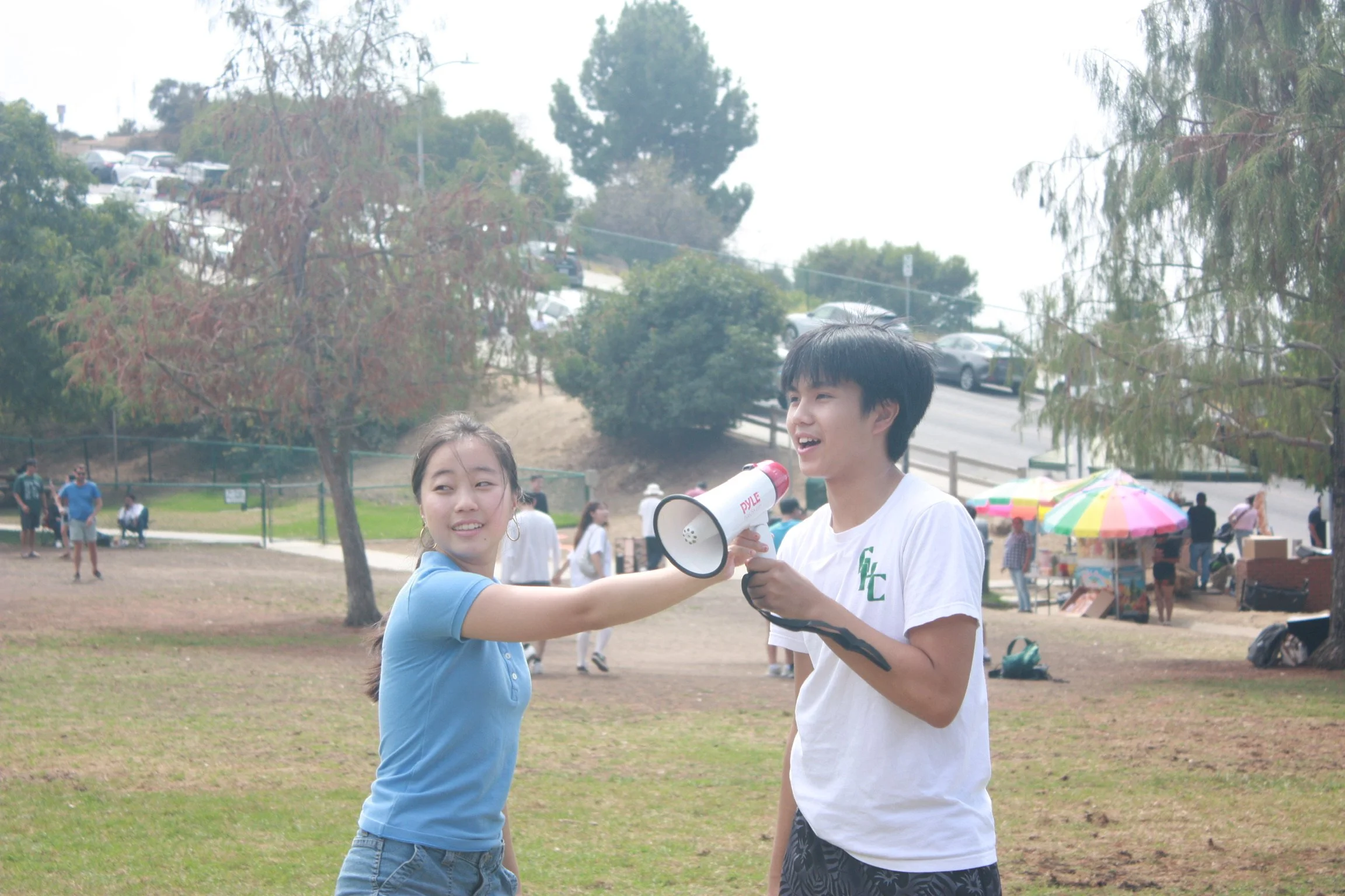 A young woman and young man are outdoors at a park. The woman is holding a megaphone and speaking into it, while the man is holding it and smiling. They are surrounded by other people, with trees, parked cars, and a vendor stand with a rainbow-colore
