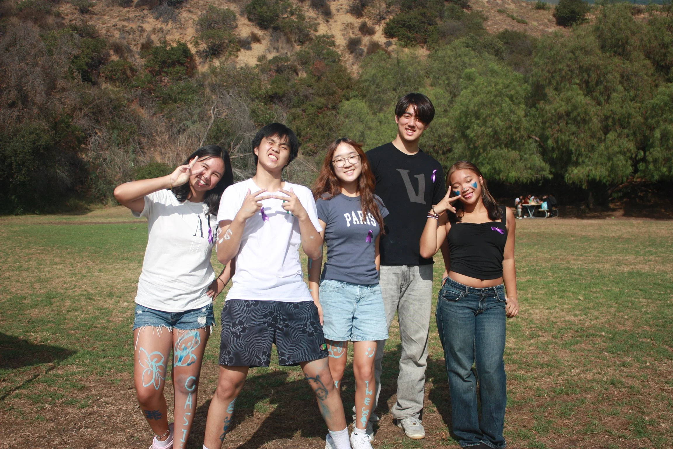 A group of five young people standing together outdoors on a sunny day, with trees and a hillside in the background. They are smiling and making peace signs or playful gestures.