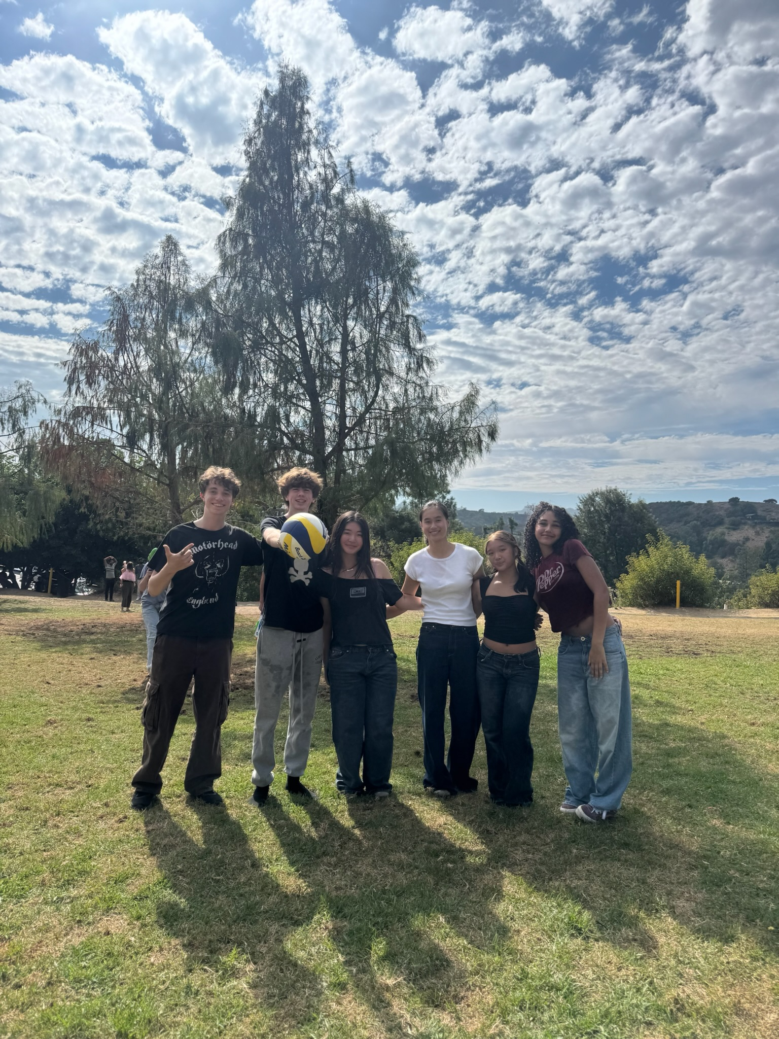 A group of seven young people standing outdoors on a grassy field, with trees and a partly cloudy sky in the background, some holding a volleyball.