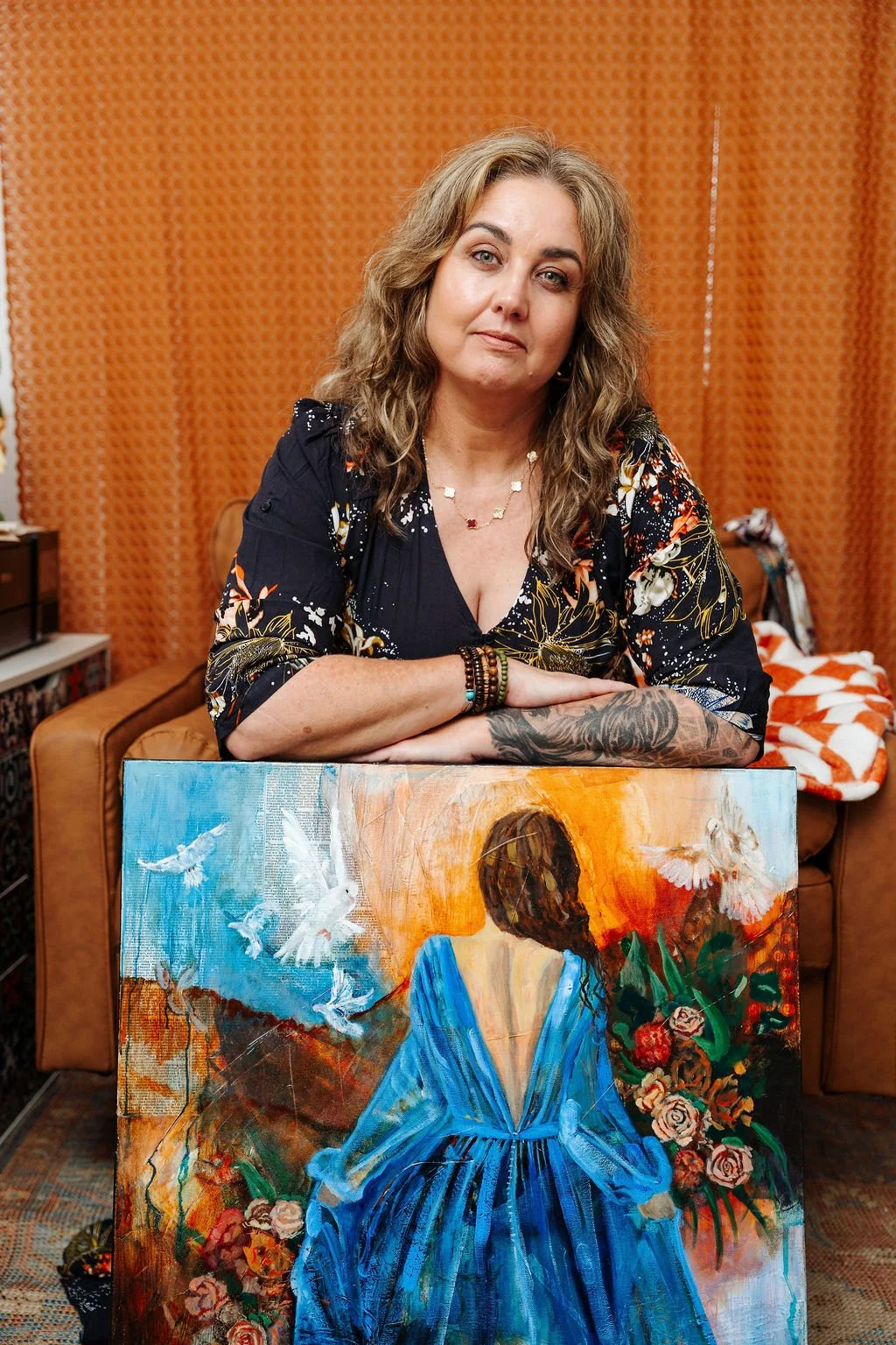 A woman with wavy light brown hair and tattoos on her arm, wearing a dark floral dress and jewelry, leaning on a colorful painting of a woman in a blue dress with a floral background, in a room with orange patterned curtains and a brown sofa.