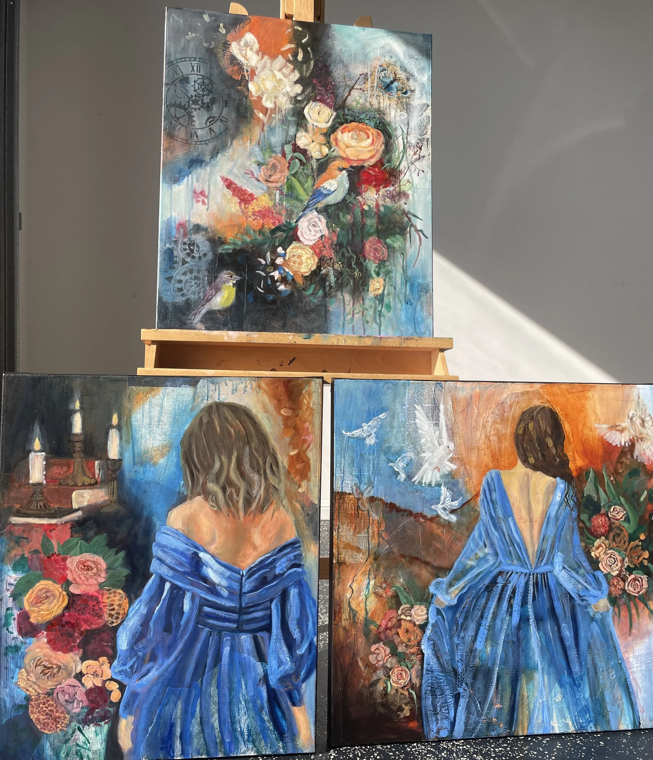 Three paintings with vibrant colors. The top painting depicts a bouquet with birds, flowers, and clock motifs. The lower left painting shows a woman in a blue dress with her back turned, surrounded by flowers and candles. The lower right painting features a woman in a blue dress with her back turned, with flying doves and flowers in the background.