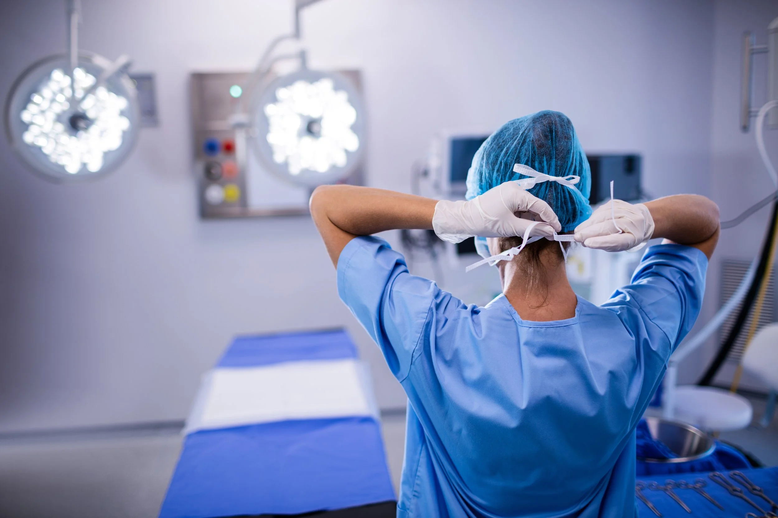 A healthcare worker in scrubs and a surgical cap preparing for surgery in an operating room with medical lights and equipment in the background.