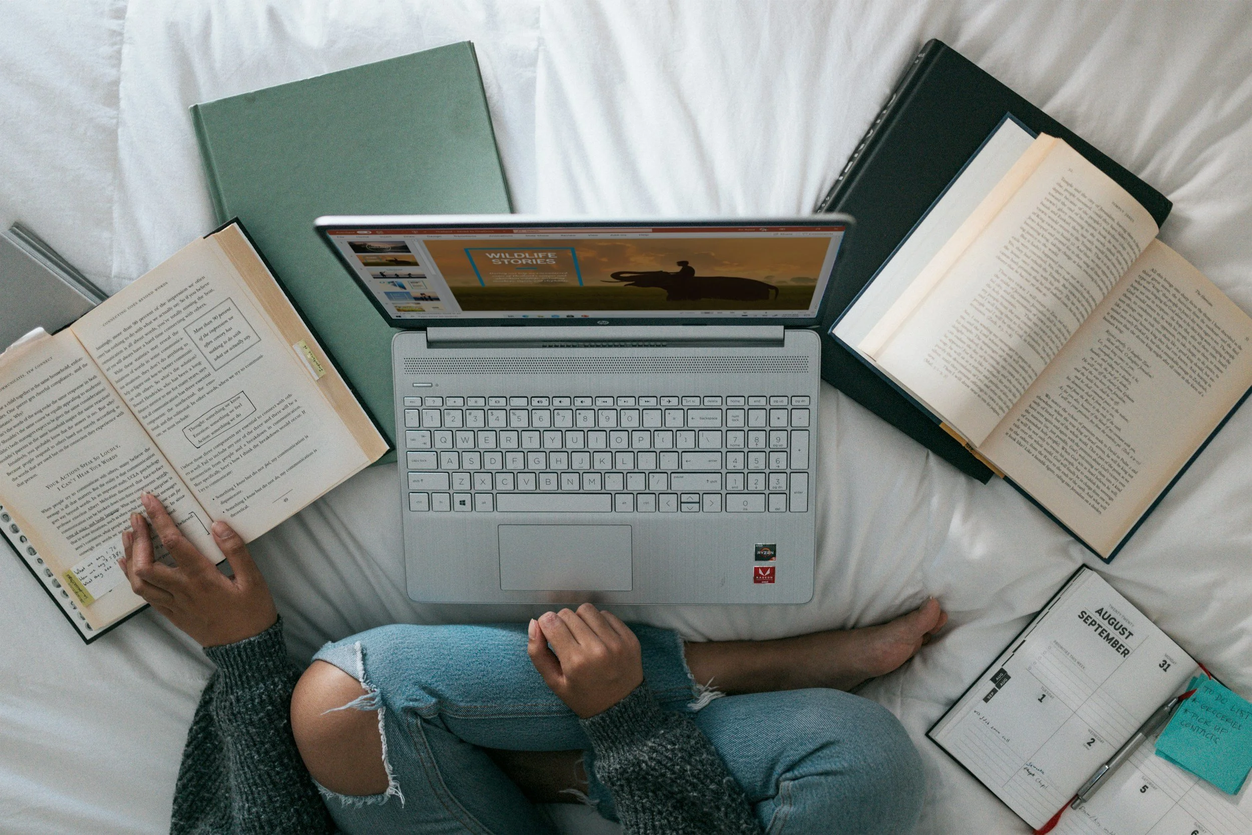 Person sitting on bed surrounded by open books, a laptop, and a calendar, studying or working.