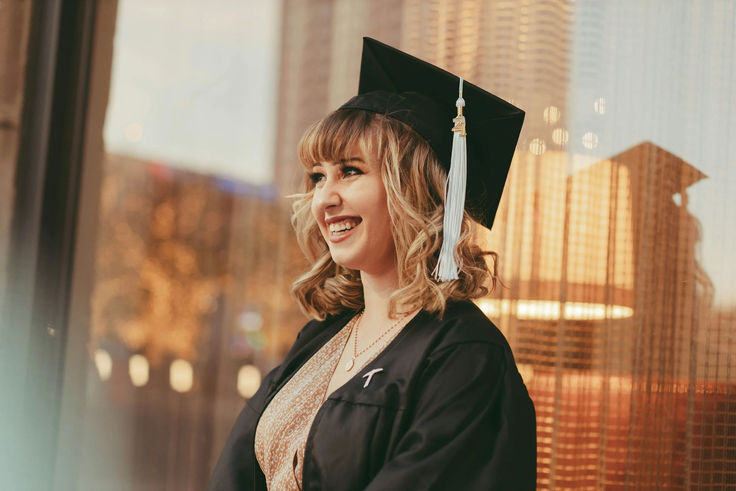 A young woman with curly blonde hair in a graduation cap and gown smiling while looking outside.
