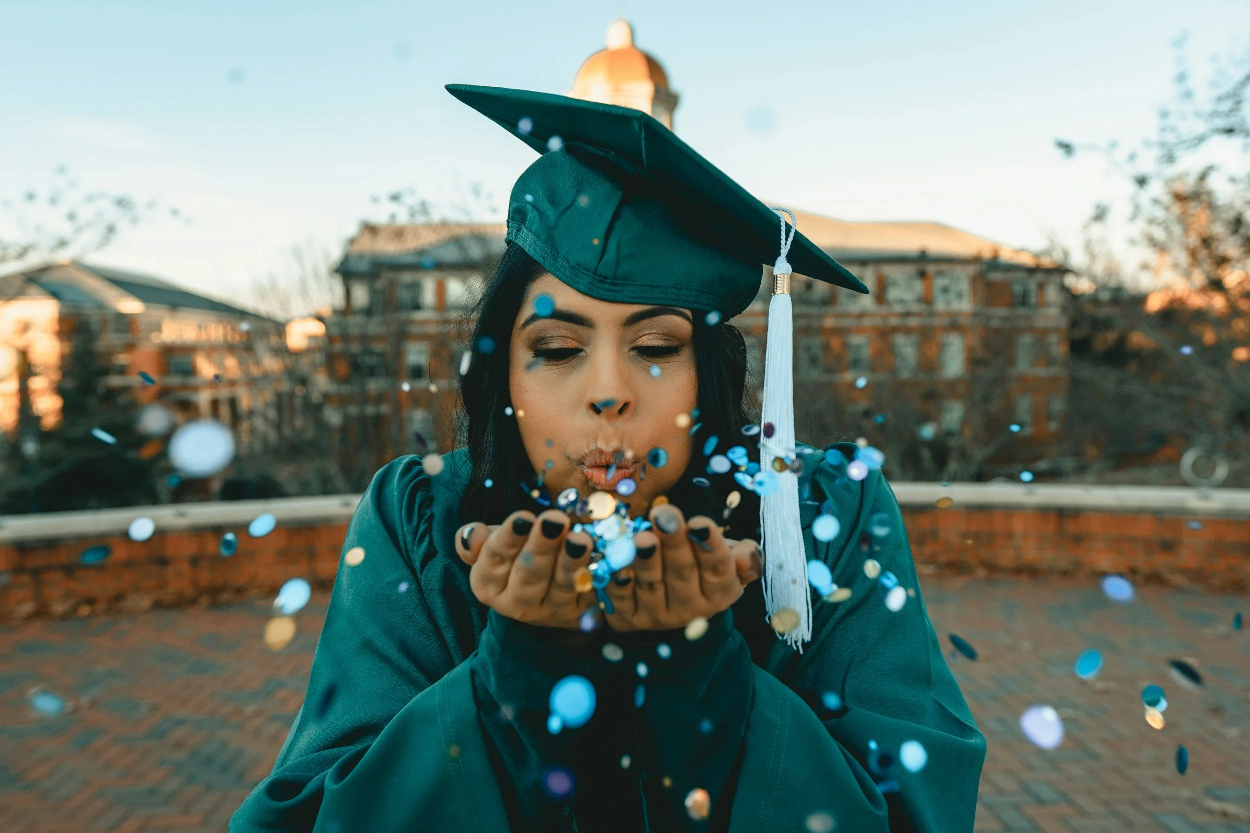 A woman in a graduation cap and gown blowing confetti on an outdoor brick courtyard with buildings in the background.