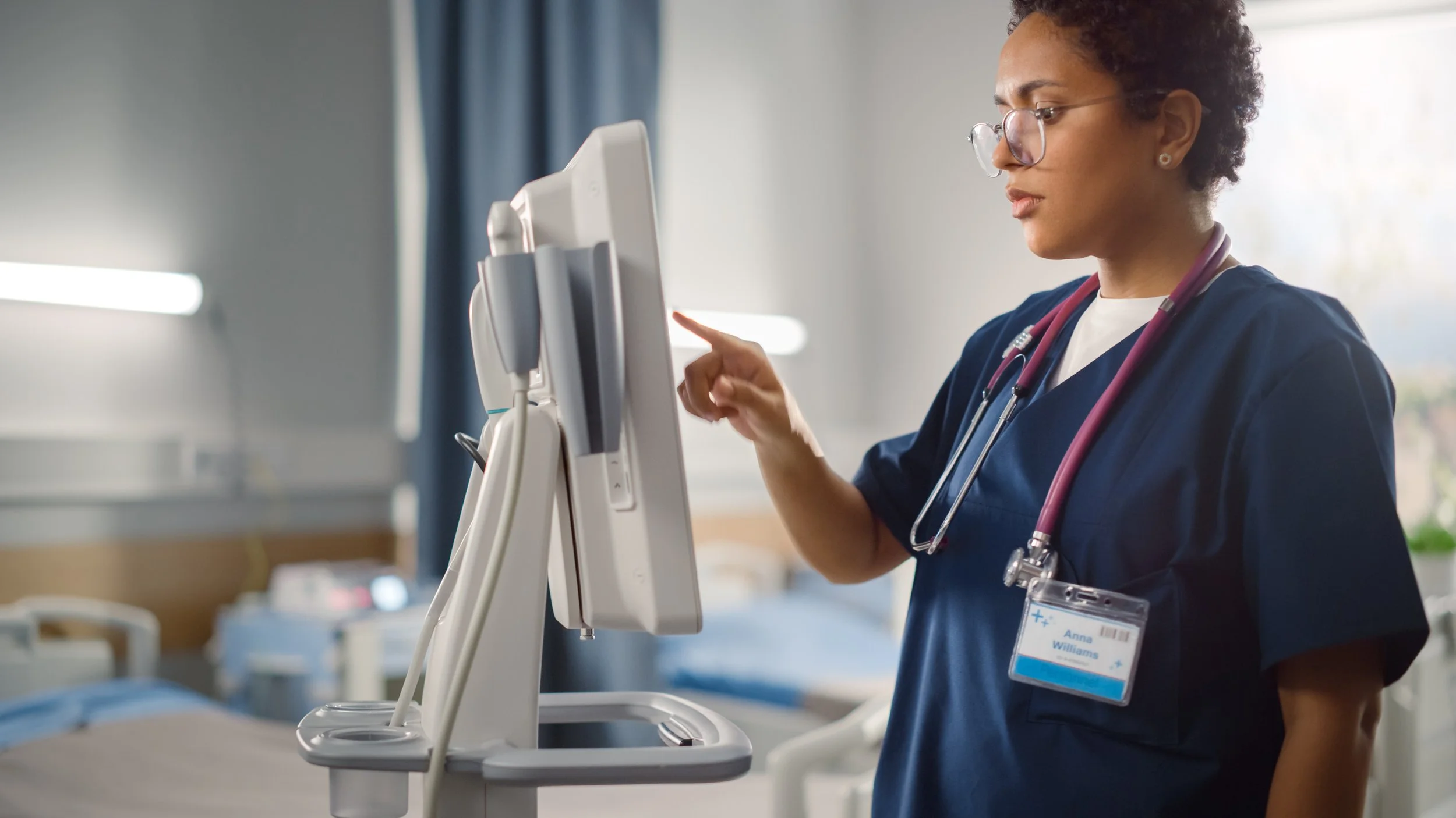 A nurse with curly hair, glasses, and a stethoscope around her neck is working on a medical monitor in a hospital room.