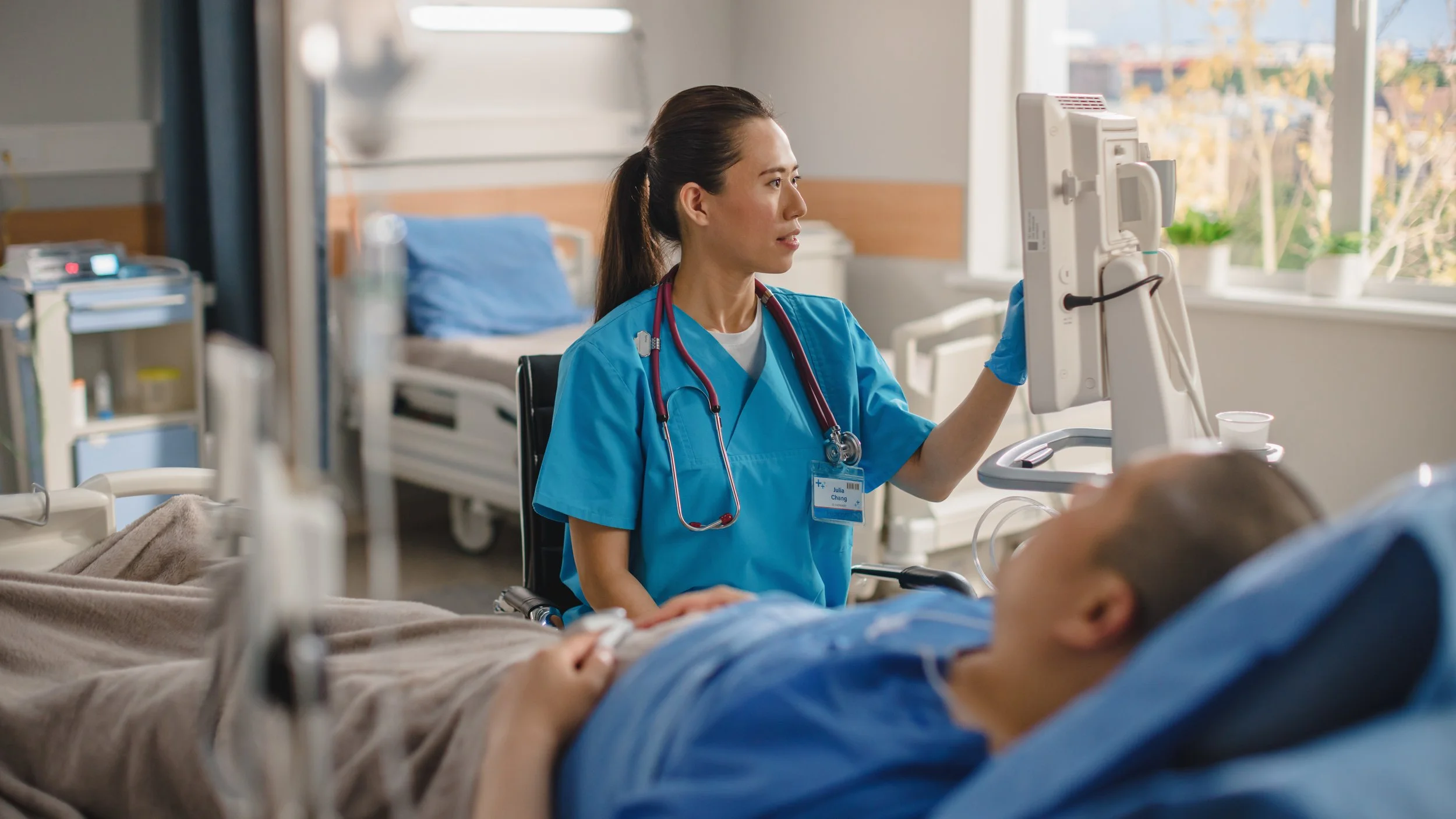 A healthcare professional in blue scrubs with a stethoscope around her neck is attending to a patient lying in a hospital bed. The nurse is looking at a monitor while the patient is in a hospital room with large windows and natural light.