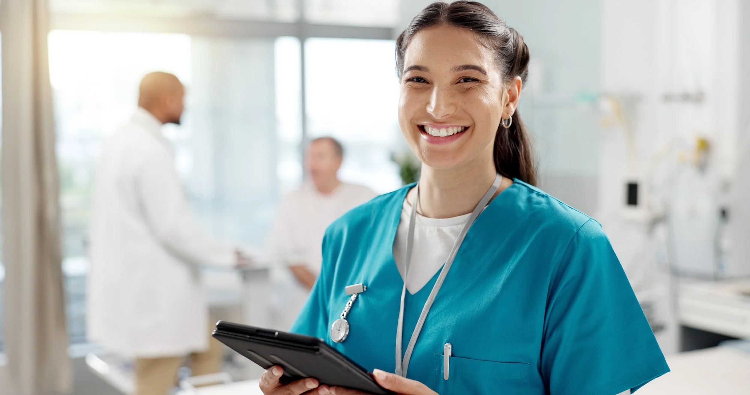 A smiling female nurse in a blue uniform holding a digital tablet, with a hospital or medical facility background showing blurred medical staff and equipment.