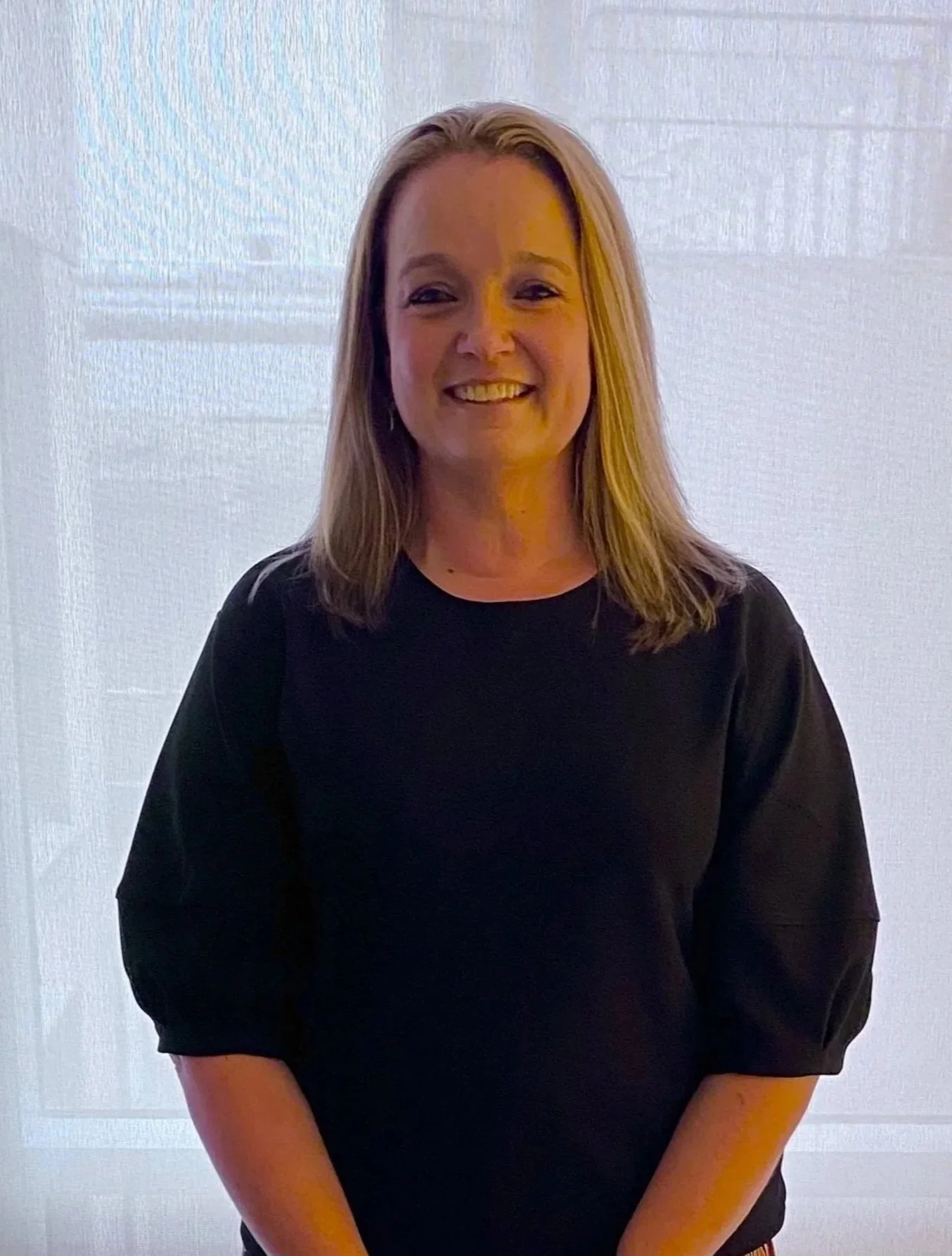 A woman with shoulder-length blonde hair smiling, wearing a black top, standing in front of a light-colored background.