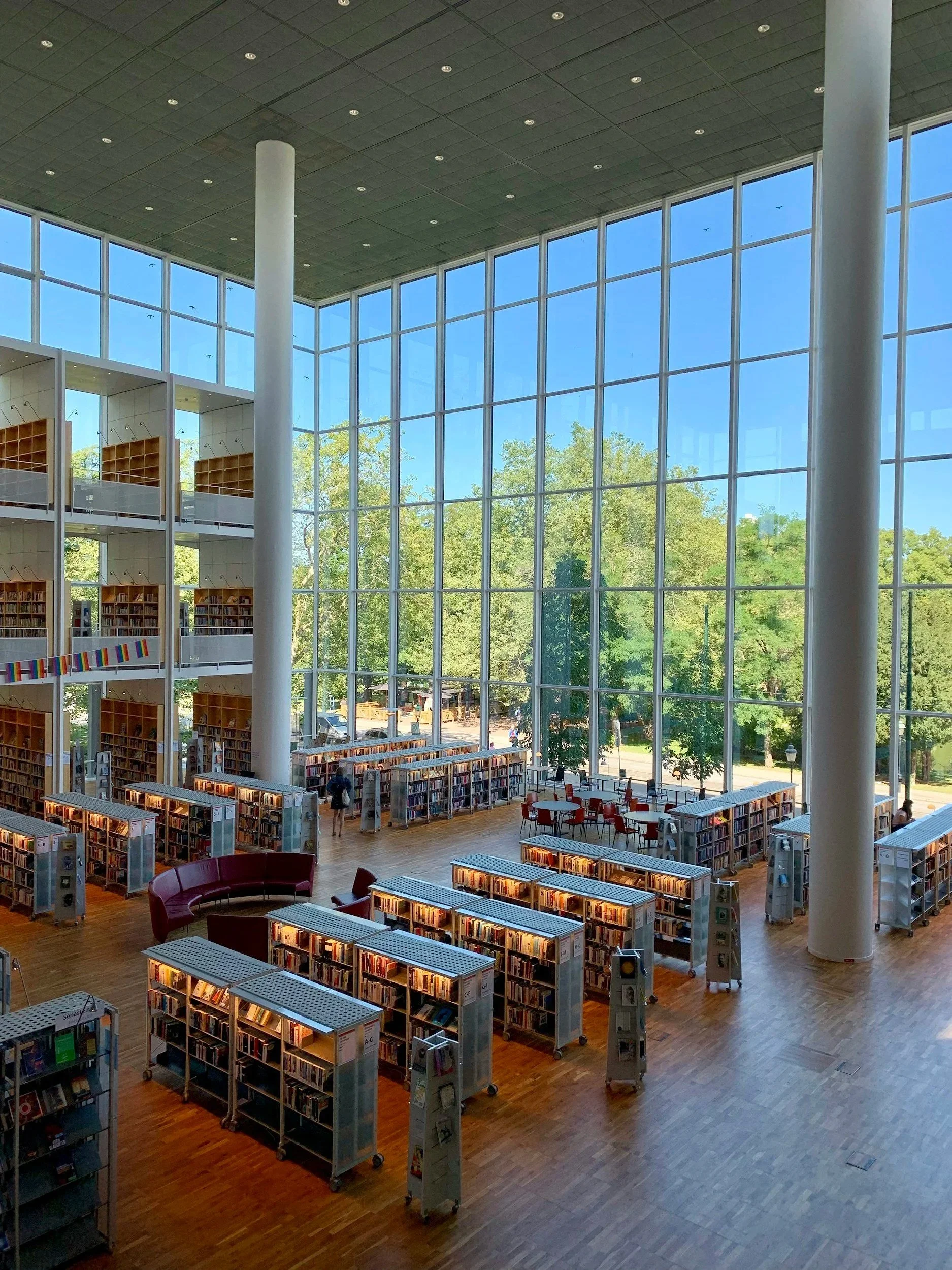 A modern library interior with tall glass windows, bookshelves, and reading areas including tables and chairs.
