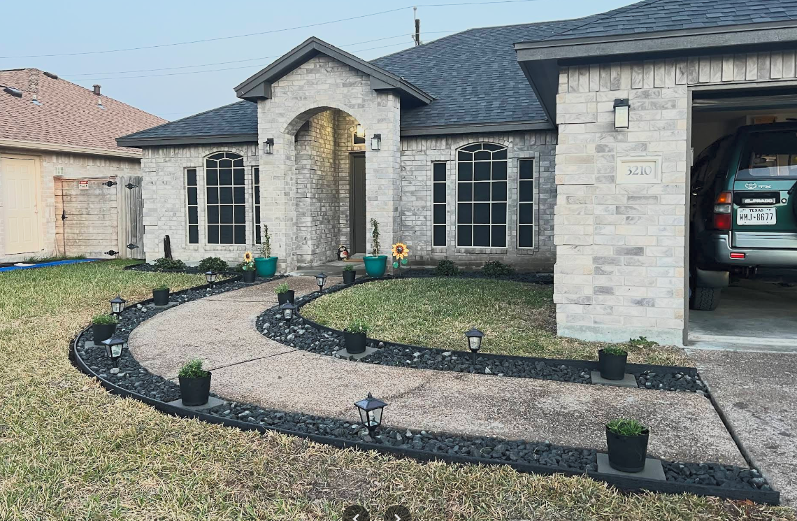 Front yard of a house with a curved concrete pathway, small landscape lights, potted plants, and a brick exterior with a garage on the right.