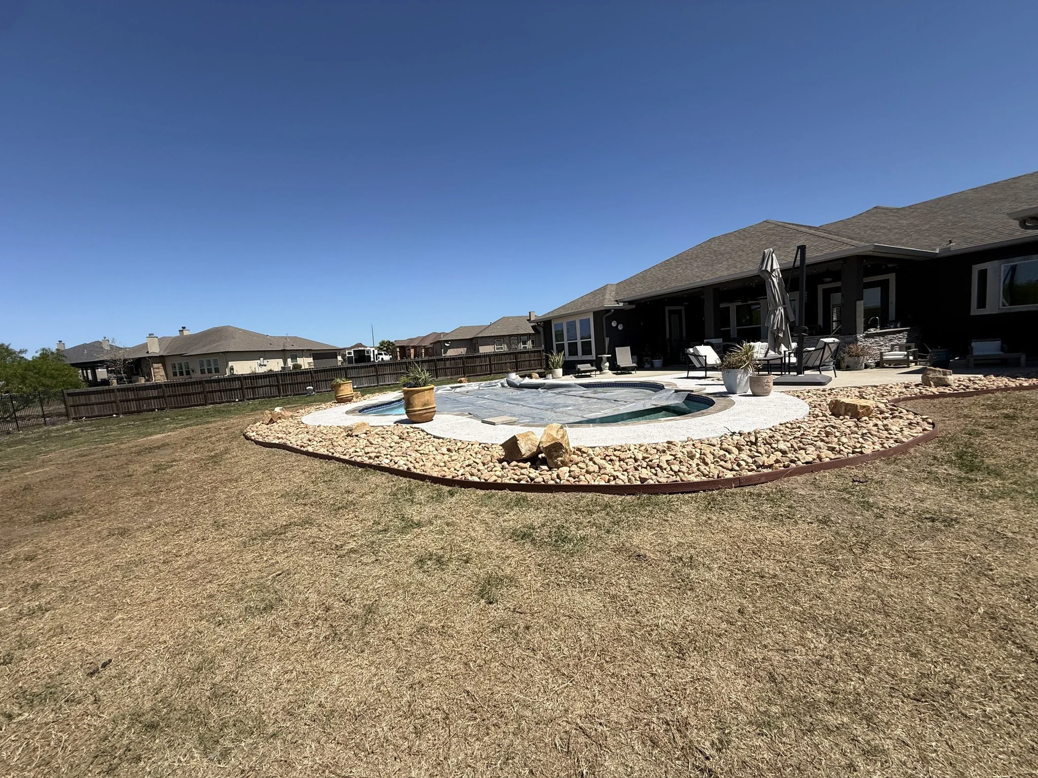 A backyard with a swimming pool surrounded by a stone patio, potted plants, outdoor furniture, and a wooden fence. Residential houses are visible in the background under a clear blue sky.