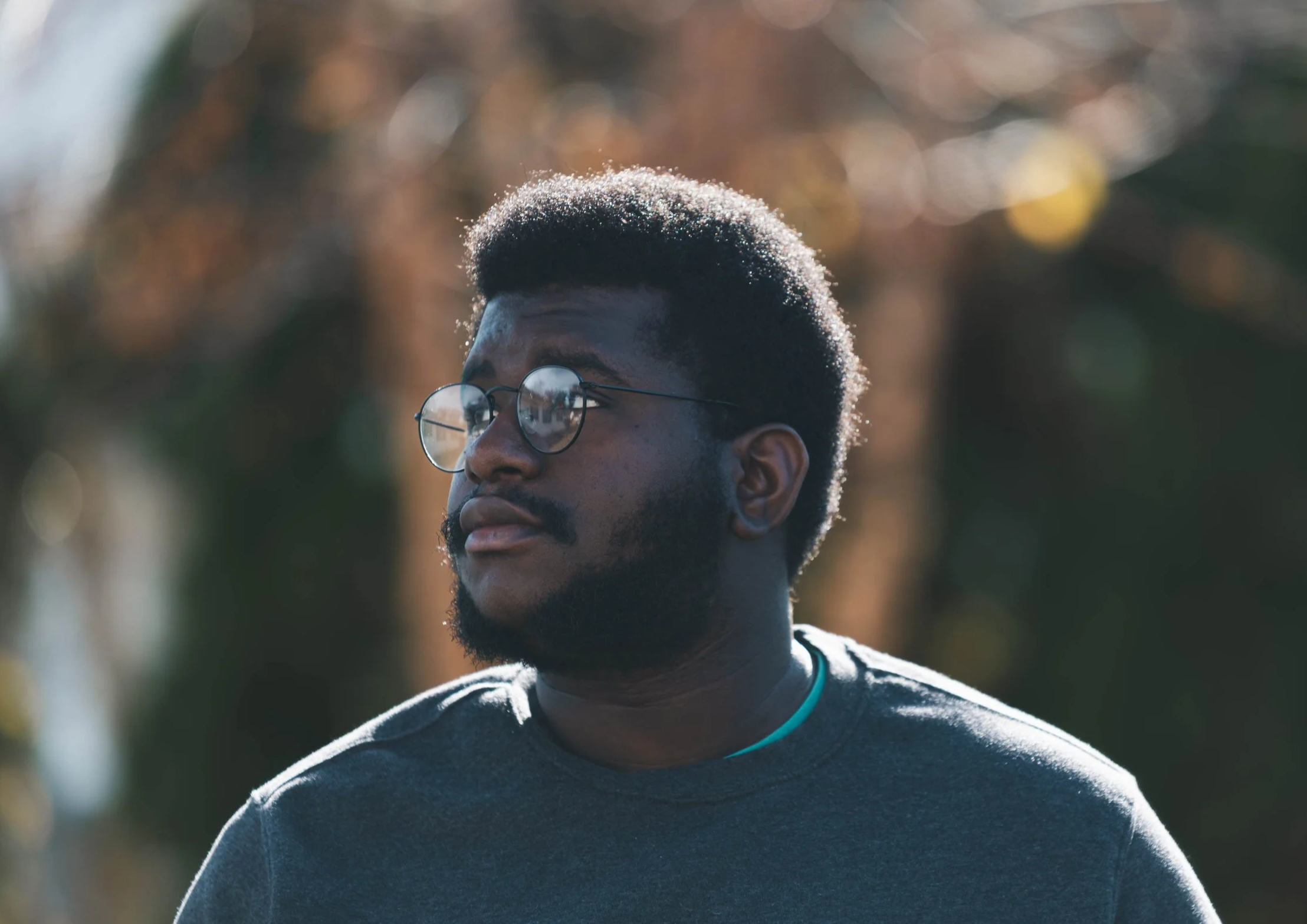 A young man with dark skin, wearing round eyeglasses and a gray T-shirt, standing outdoors with blurred trees in the background, bathed in warm sunlight.