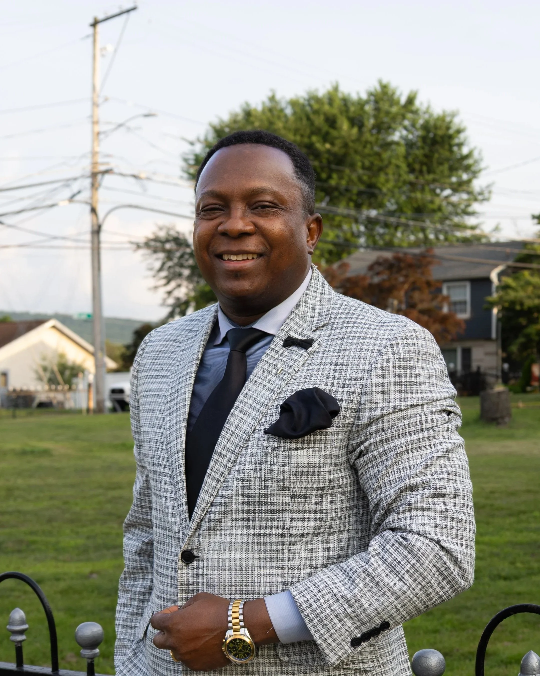 A man dressed in a checkered blazer, blue shirt, and black tie, standing outdoors in a grassy area during daylight. He is smiling and wearing a gold watch and a black pocket square. There are trees, houses, and power lines in the background.