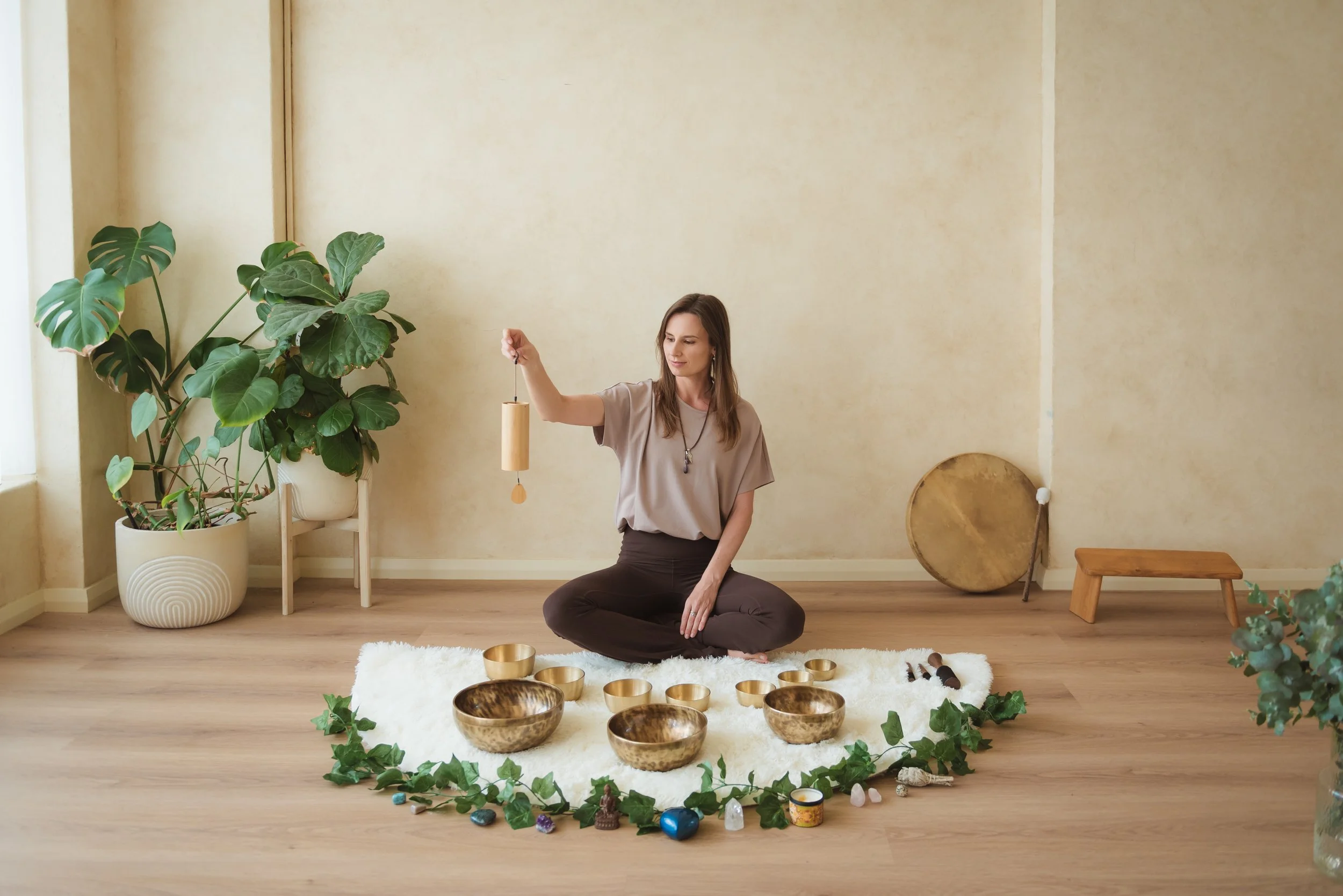 A woman sitting cross-legged on a white rug. There are singing bowls in front of her, decorated with green ivy. The room has light-colored walls, a wooden floor, a potted plant on the left, and a large drum leaning against the wall on the right.