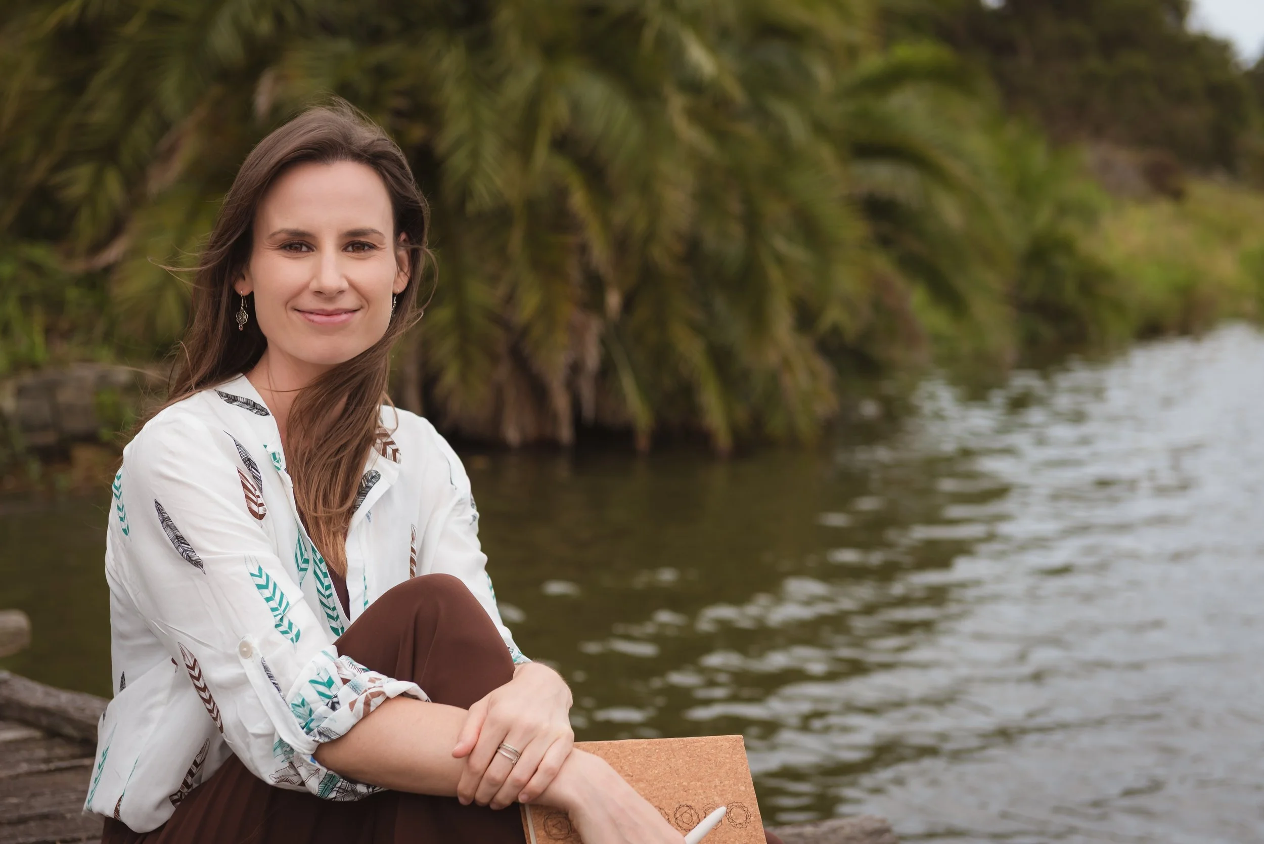 A woman sitting outdoors near a body of water, surrounded by lush greenery, smiling at the camera.