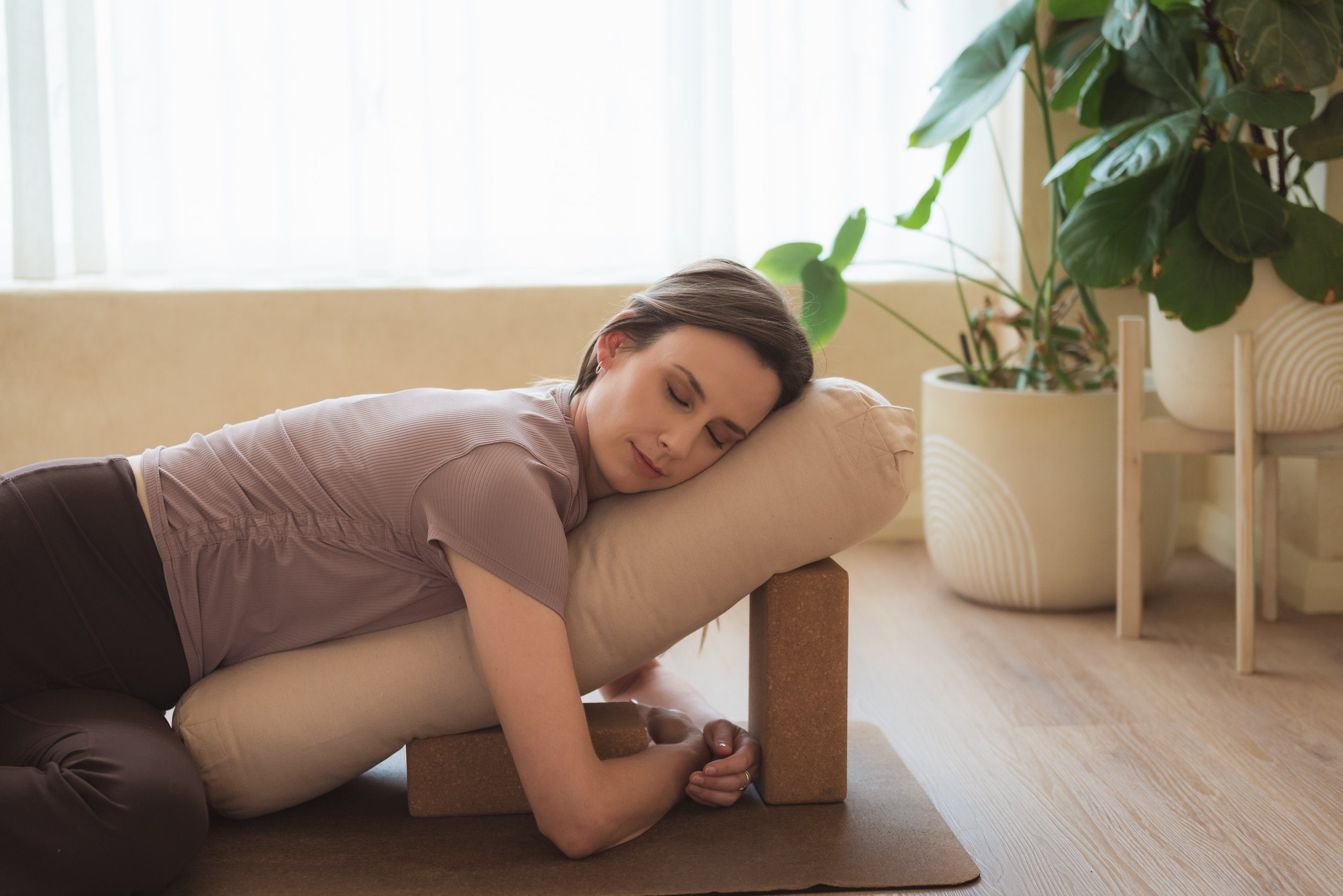 Woman relaxing with eyes closed, lying on a pillow supported by yoga blocks on the floor in a cozy room with large windows and potted plants.