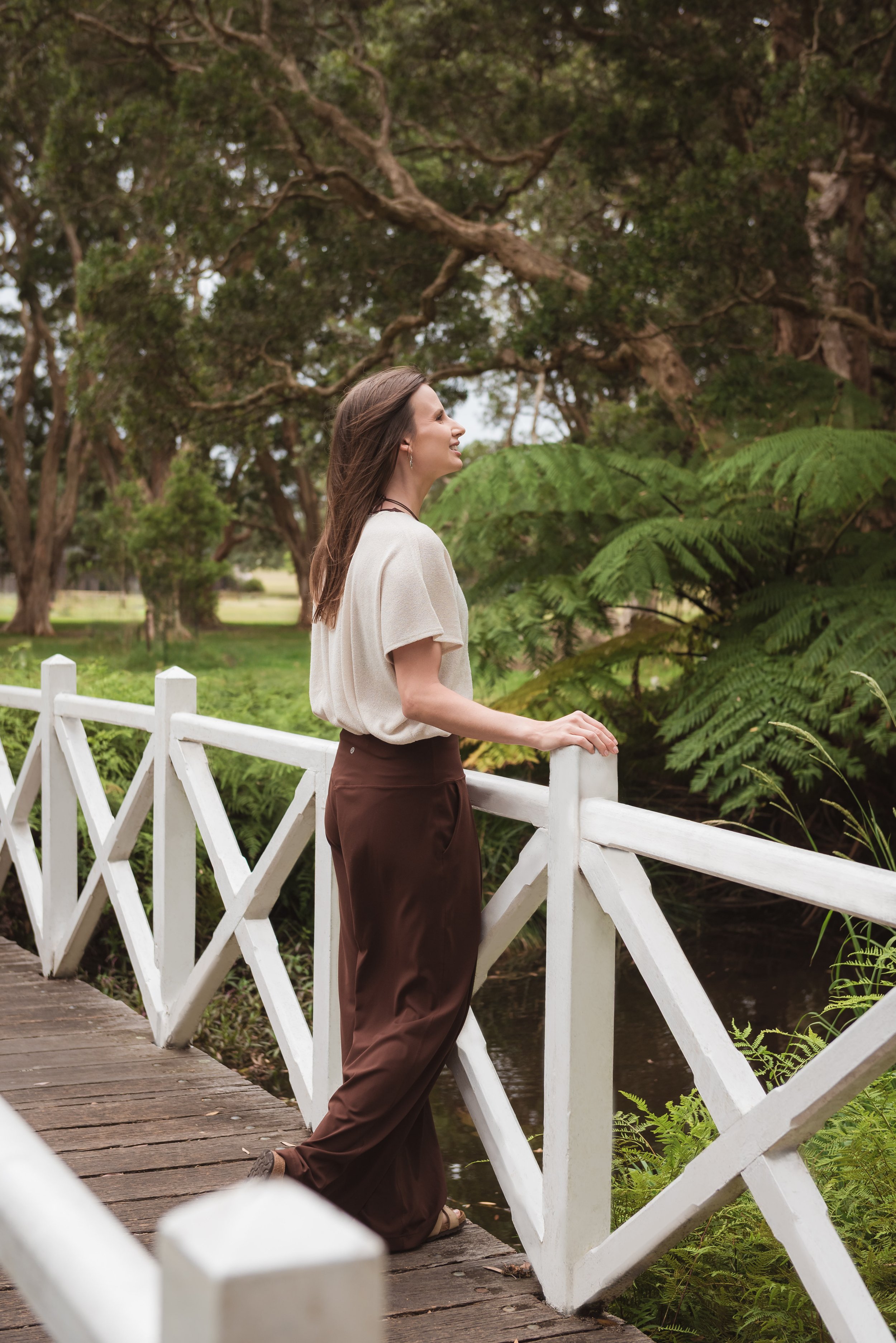 A woman standing on a wooden bridge, leaning on the white railing, smiling and looking to her right, surrounded by lush green trees and foliage.