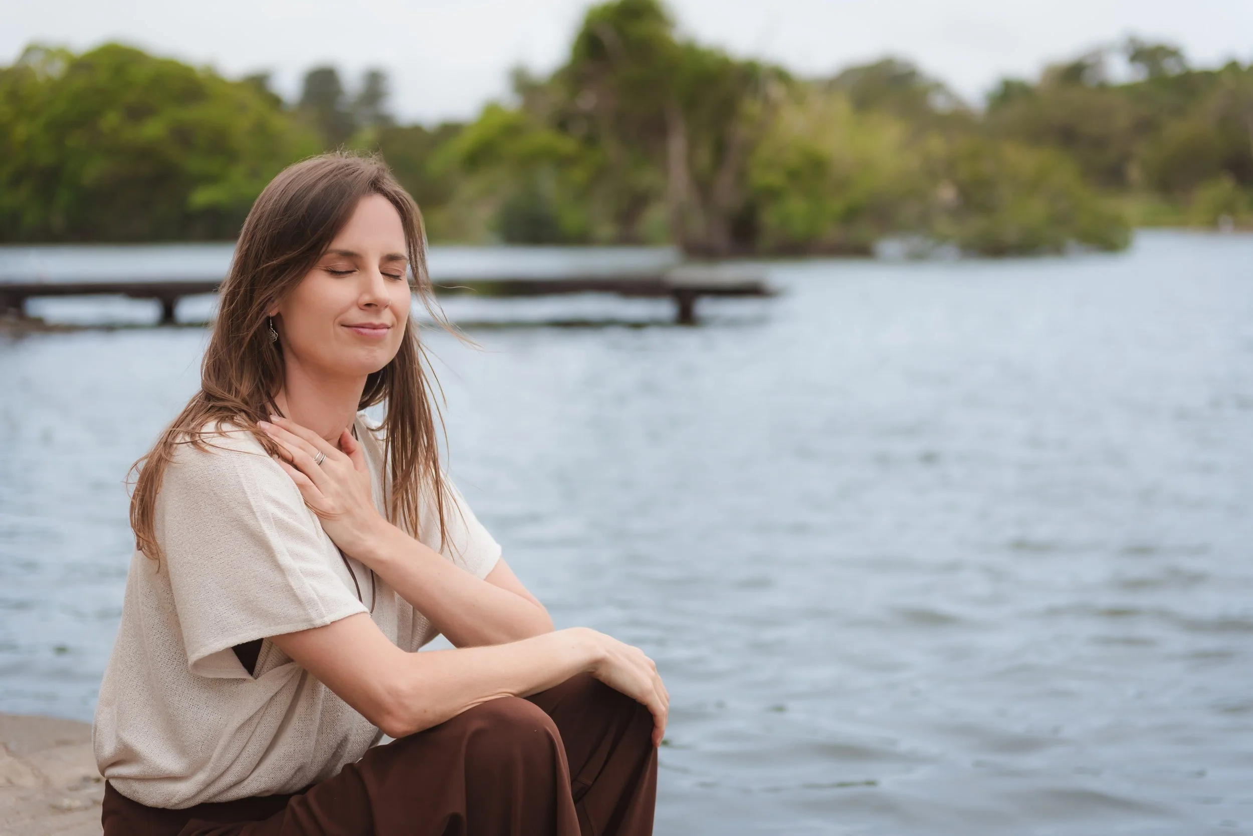 A woman sitting by a body of water with her eyes closed and her hand on her heart, appearing to be in a moment of emotional reflection or meditation, with trees in the background.