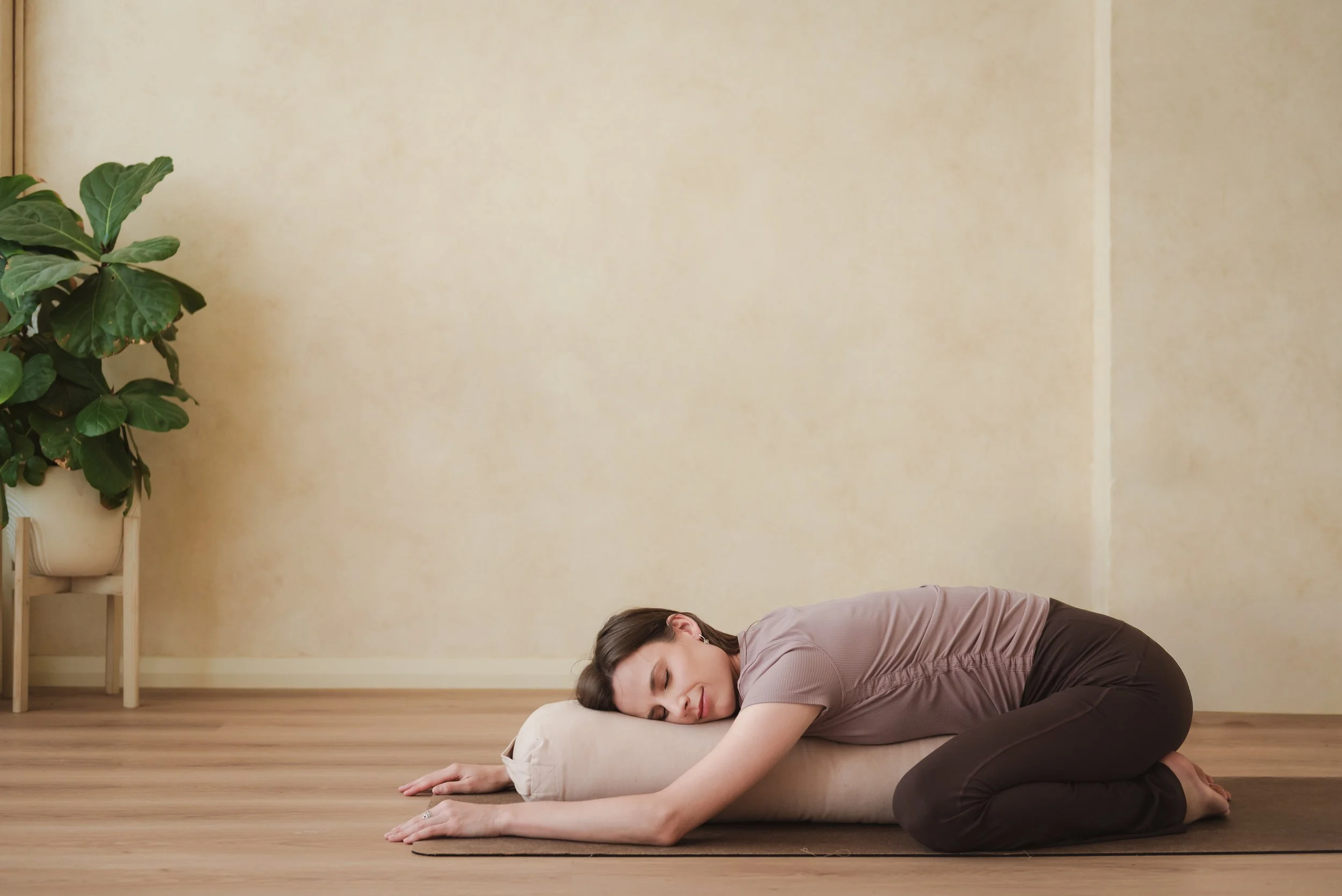 A woman practicing yin yoga on a pillow and blanket in a peaceful room with a large green plant.