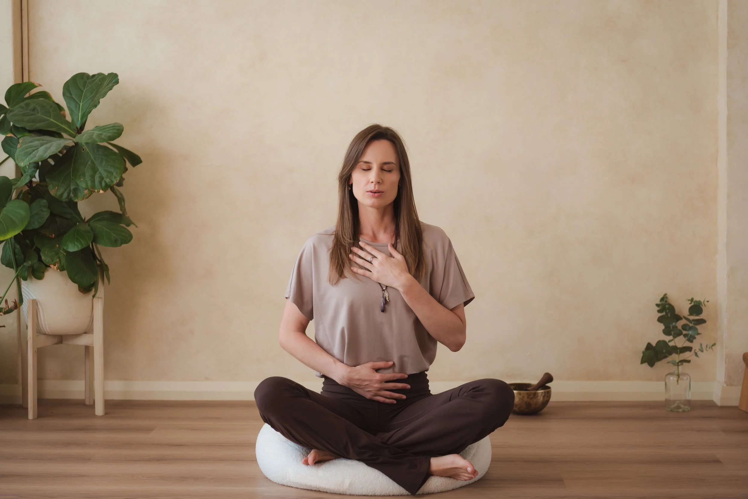 A woman practicing meditation and mindfulness seated cross-legged on a cushion indoors with plants and a singing bowl around her.