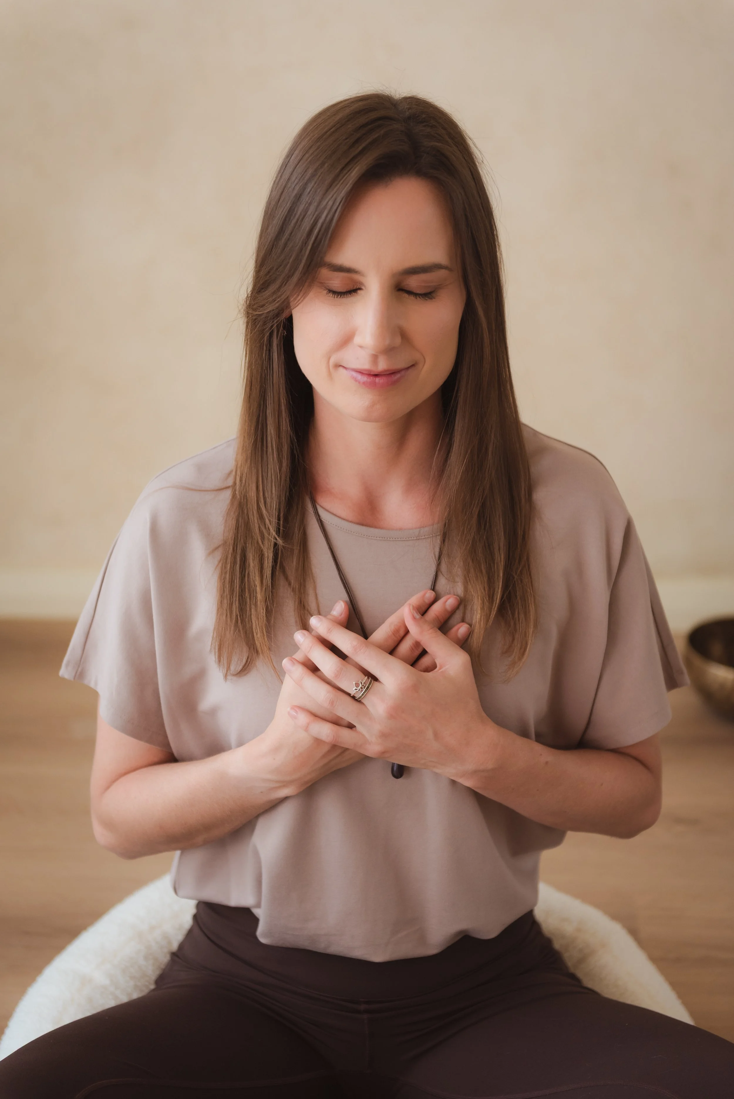 A woman with long brown hair and closed eyes sits cross-legged and places her hands on her chest in a peaceful pose.