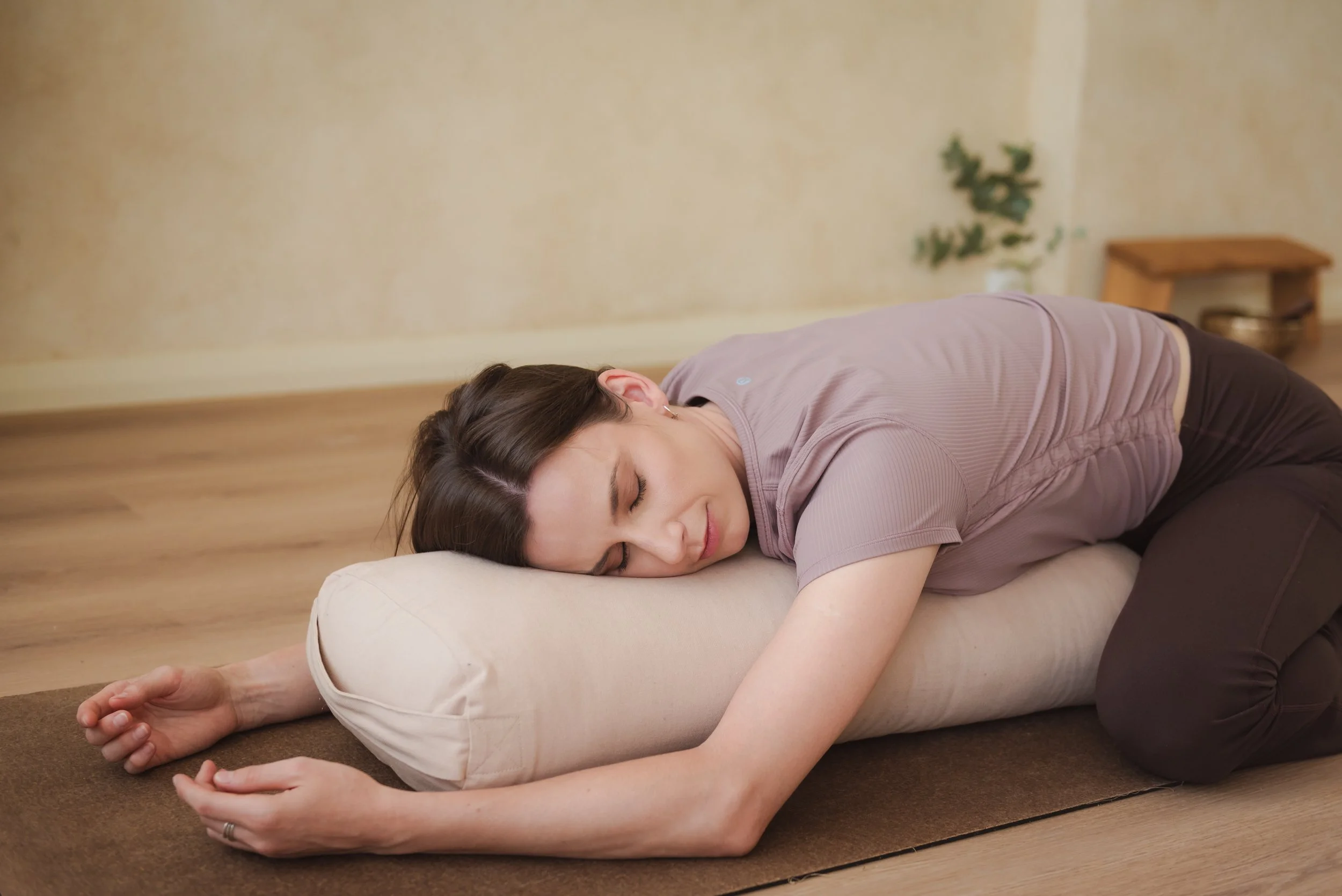 A woman practicing child's pose yoga on a cushion in a room with wooden floors and beige walls.