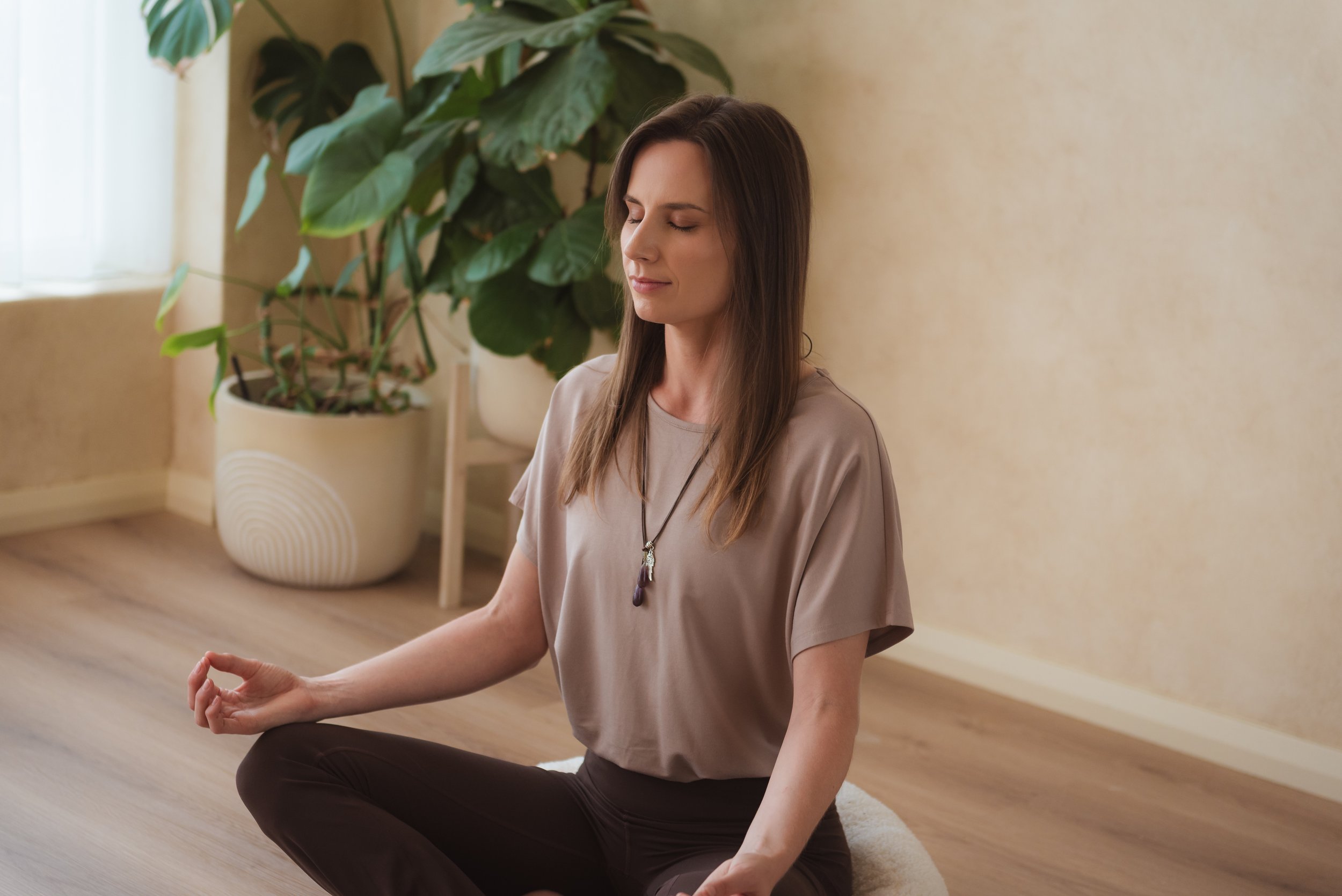 A woman practicing meditation indoors, sitting in a cross-legged position on a cushion with her eyes closed, near a large houseplant.