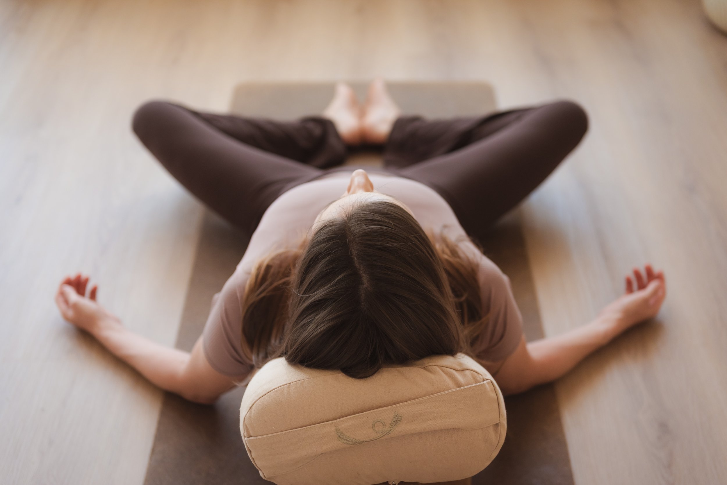 Top view of a woman practicing yin yoga on a mat, seated in a wide-legged stretch with her head resting on a bolster.