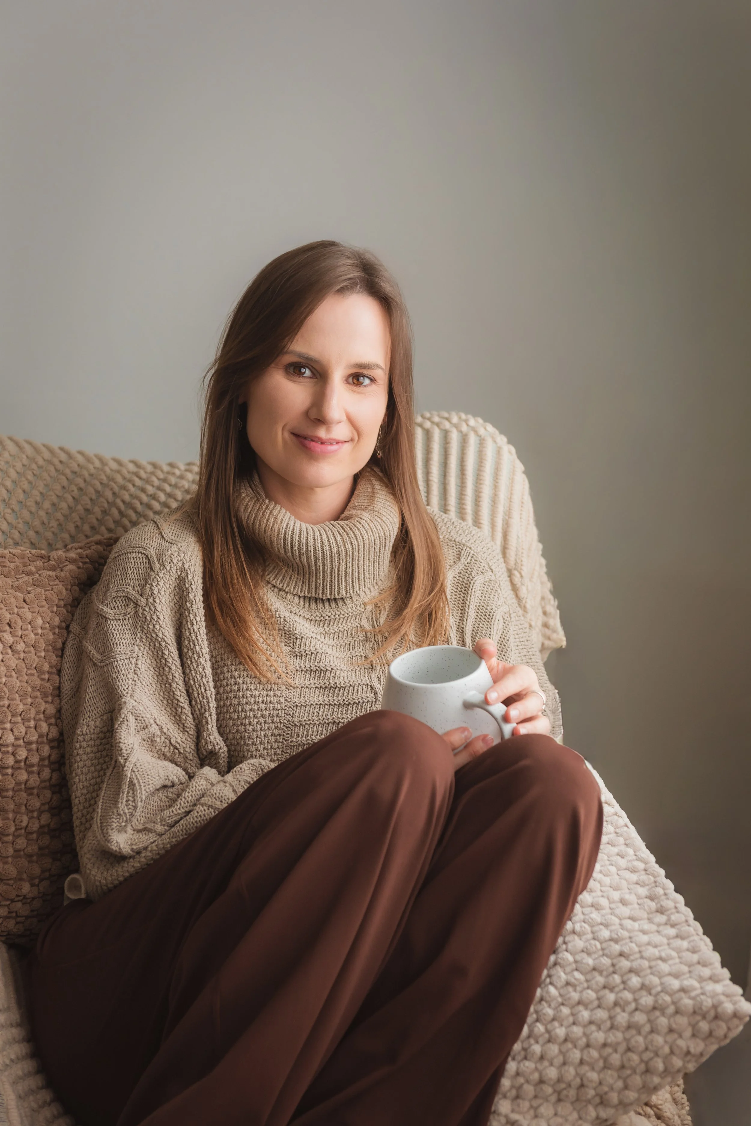 A young woman with brown hair, wearing a beige turtleneck sweater, sitting on a couch with textured pillows, holding a white ceramic mug and smiling at the camera.
