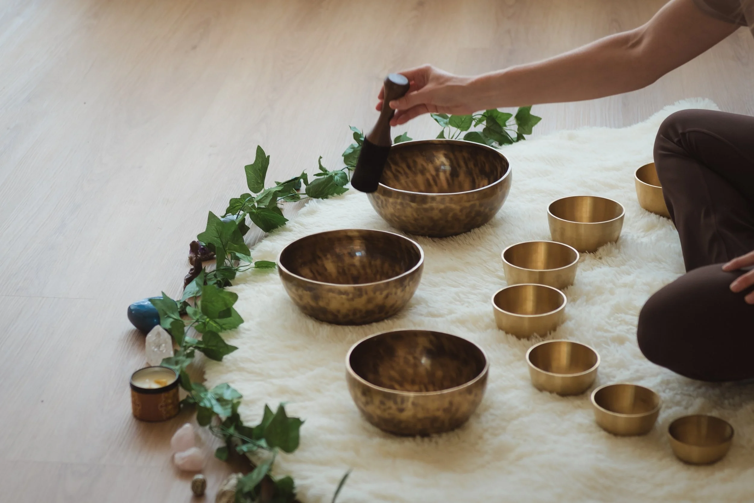 Person playing singing bowls and chimes outdoors on a white fur rug, surrounded by green ivy and crystals.