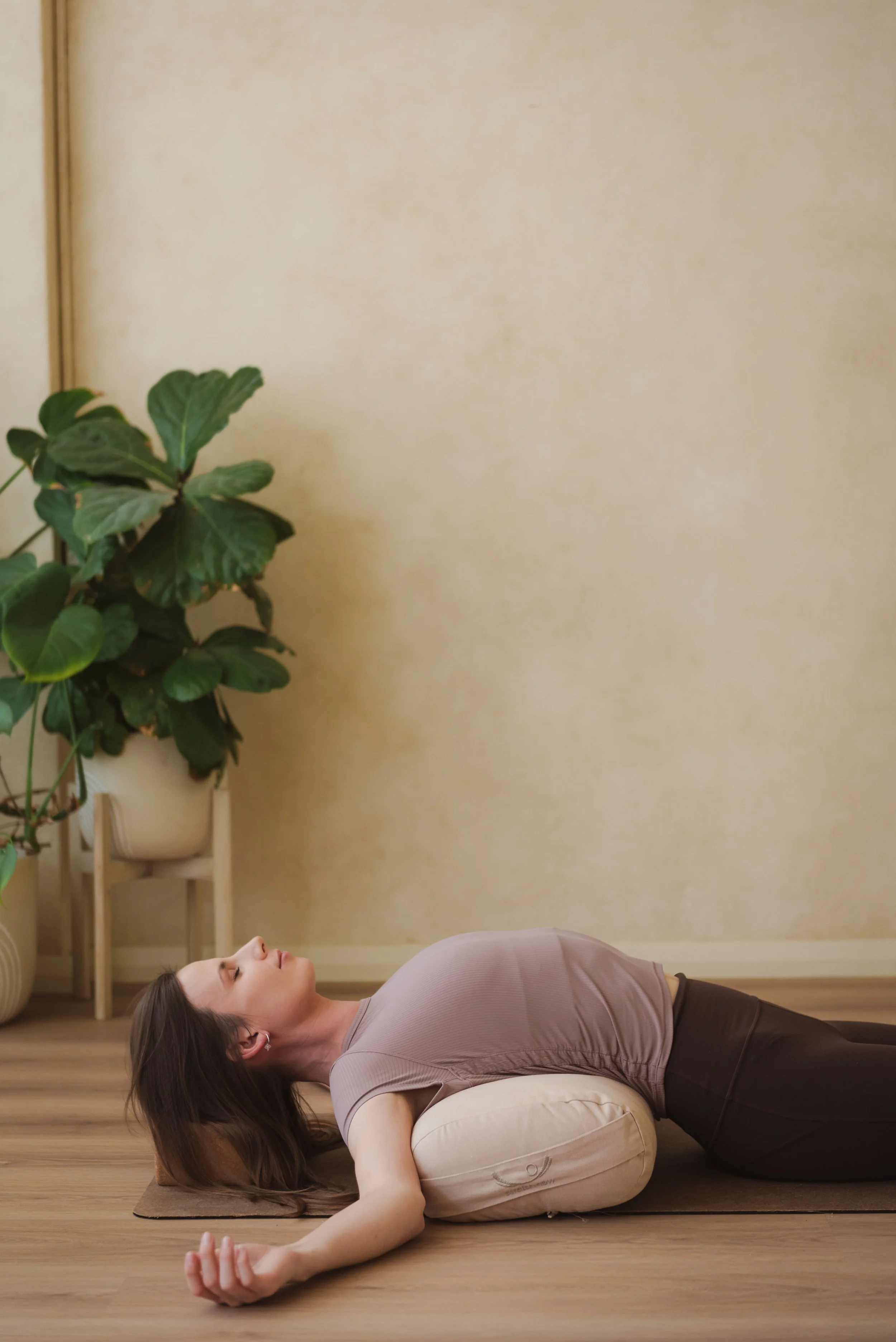 A woman practicing restorative yin yoga, lying on a mat with a bolster support under her back, in a room with wooden floors and a large green plant in the background.
