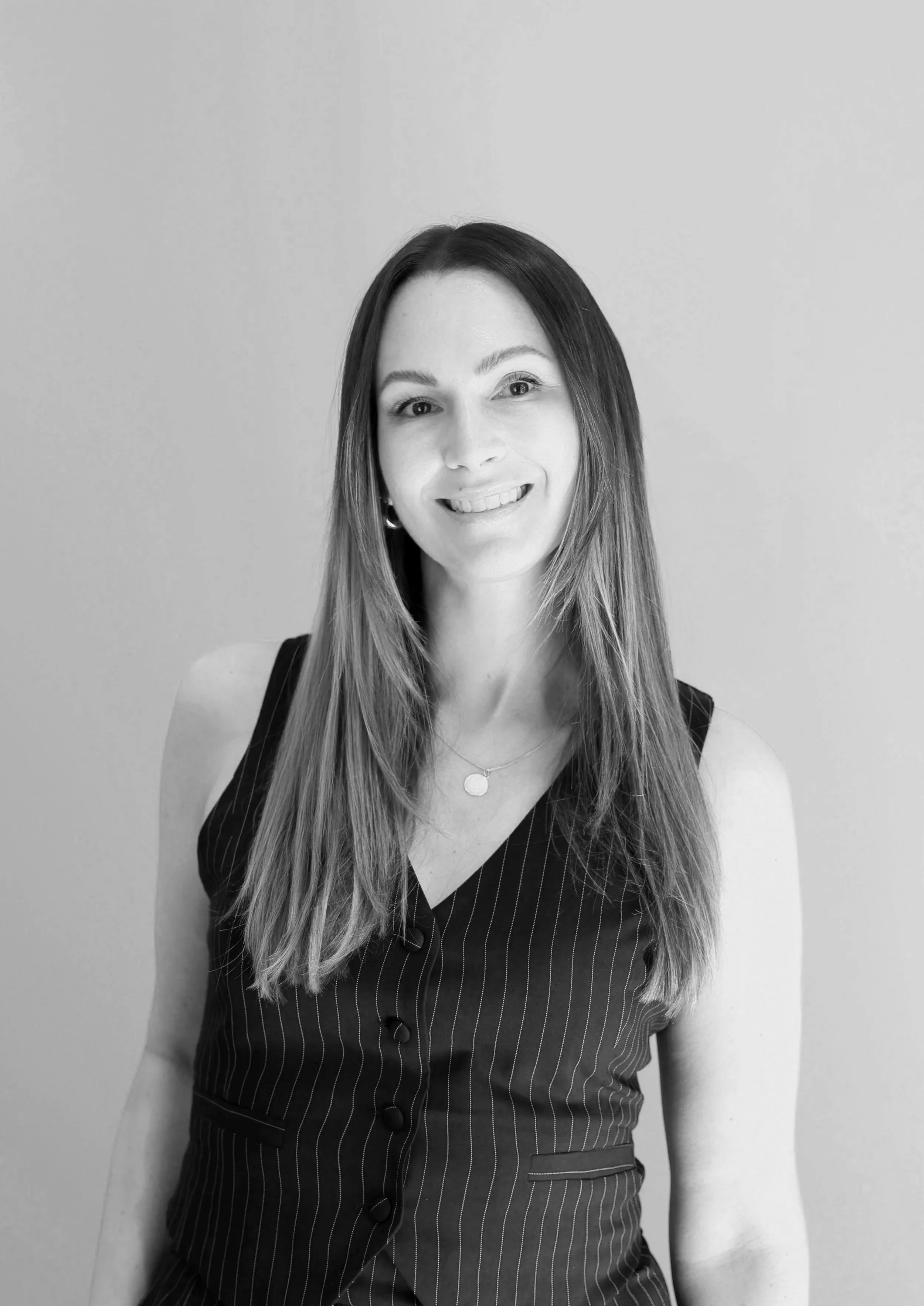 Black and white photo of a smiling woman with long straight hair, wearing a pinstriped sleeveless dress and a necklace, standing against a plain wall.