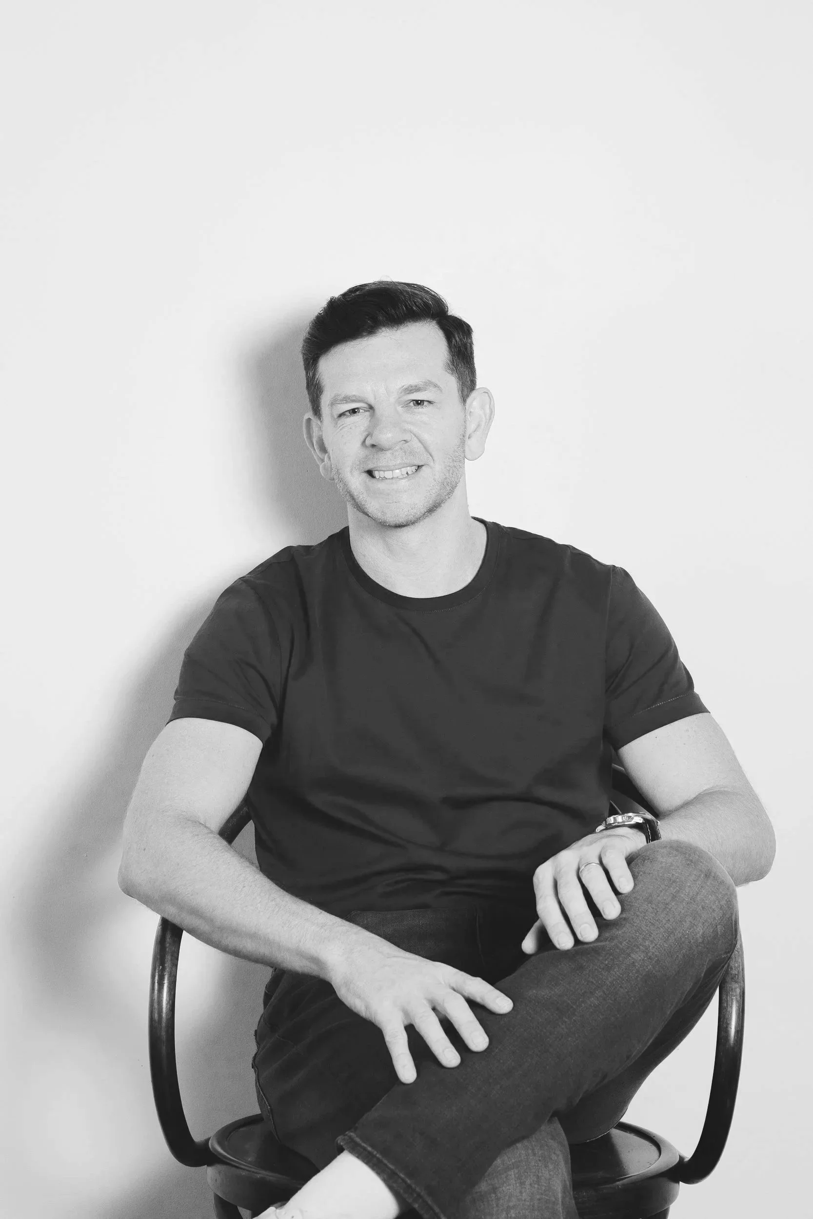 A black and white photo of a smiling man sitting on a chair with his arms crossed, wearing a black t-shirt and jeans, with a plain wall in the background.