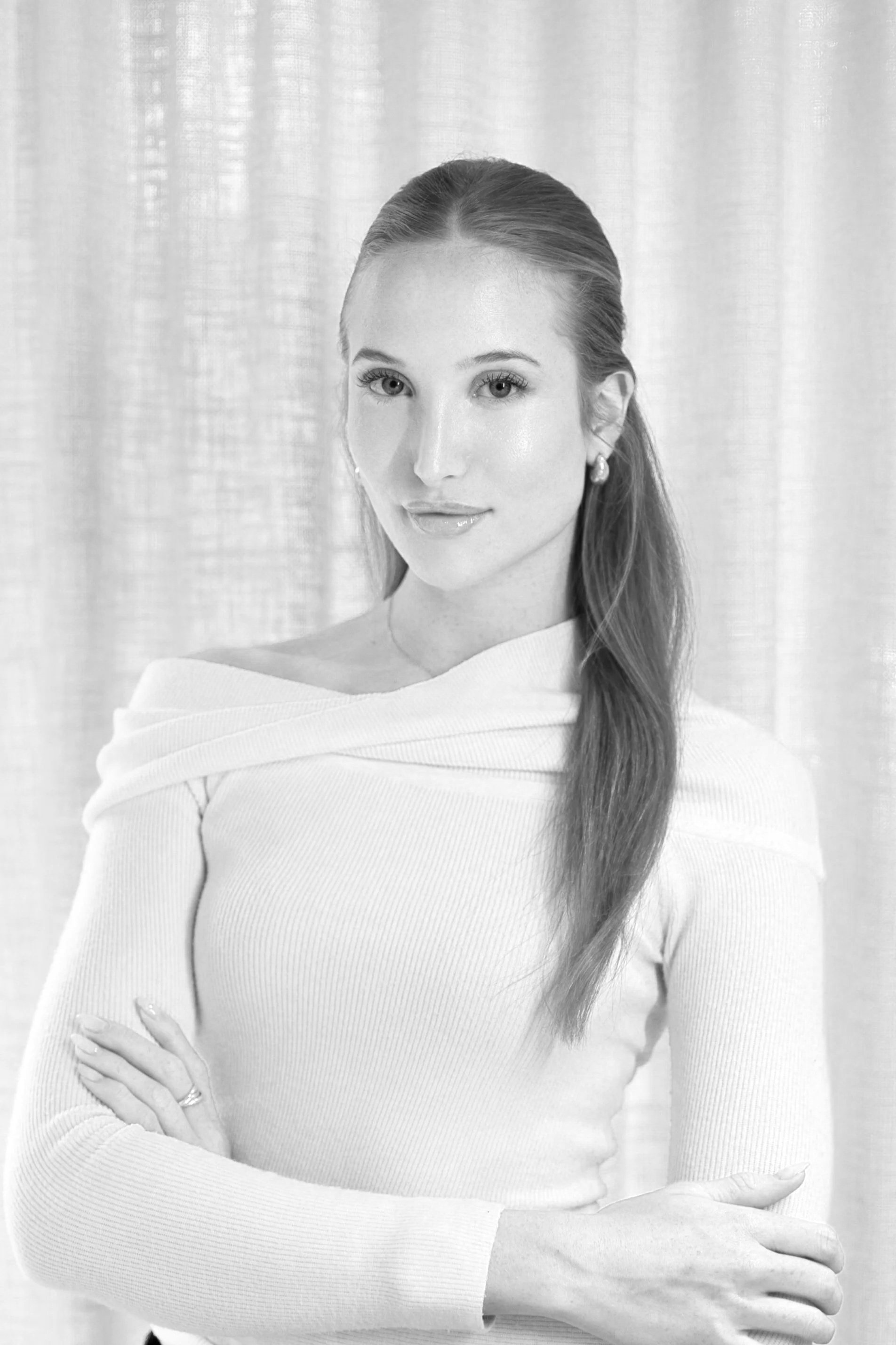 A woman with long hair wearing a light-colored off-the-shoulder sweater, standing with crossed arms in front of a textured curtain background.