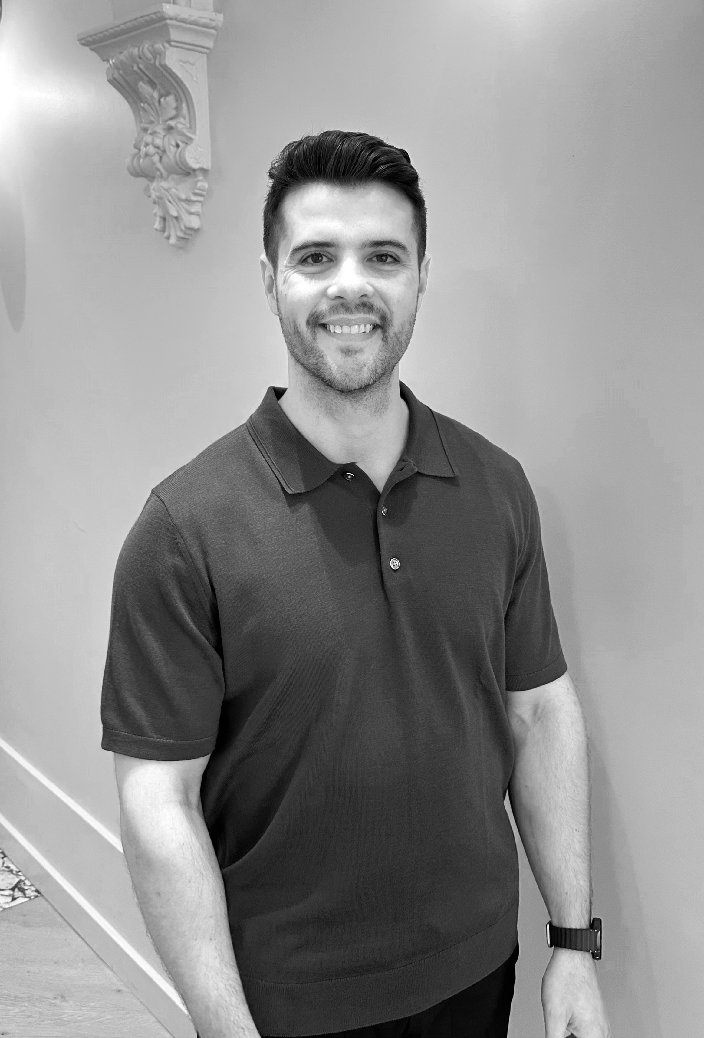 A young man with dark hair and a beard, smiling, wearing a short-sleeved polo shirt and a smartwatch on his left wrist, standing indoors in front of a plain wall with decorative molding.