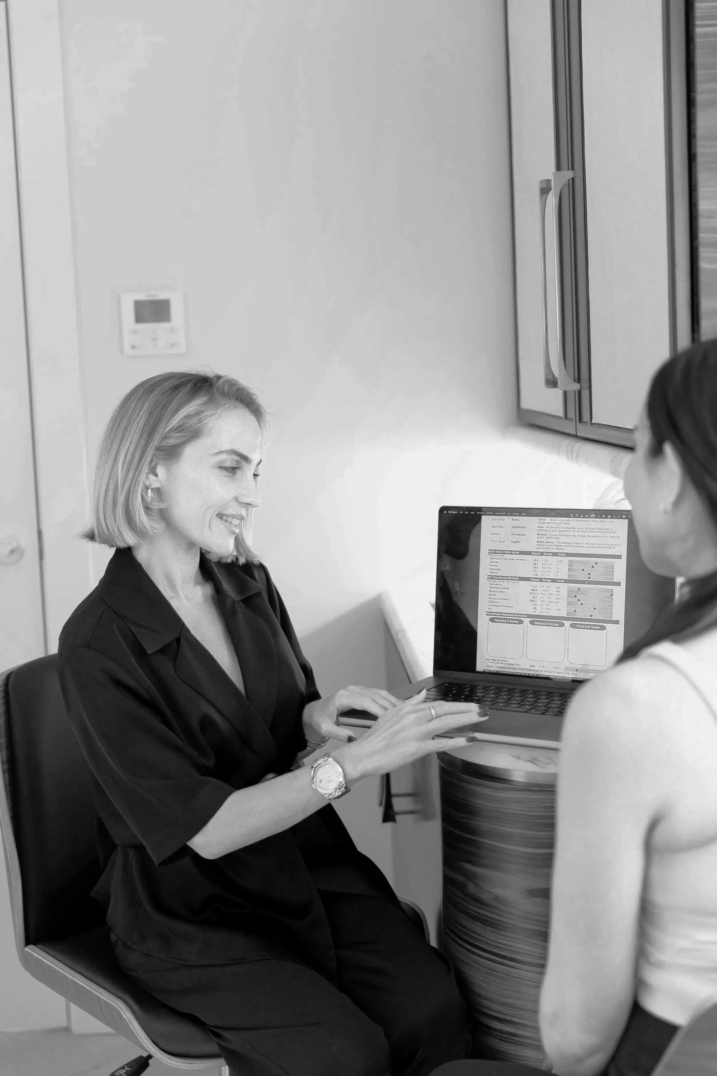 Two women sitting at a table, one showing a laptop screen to the other in an office setting.
